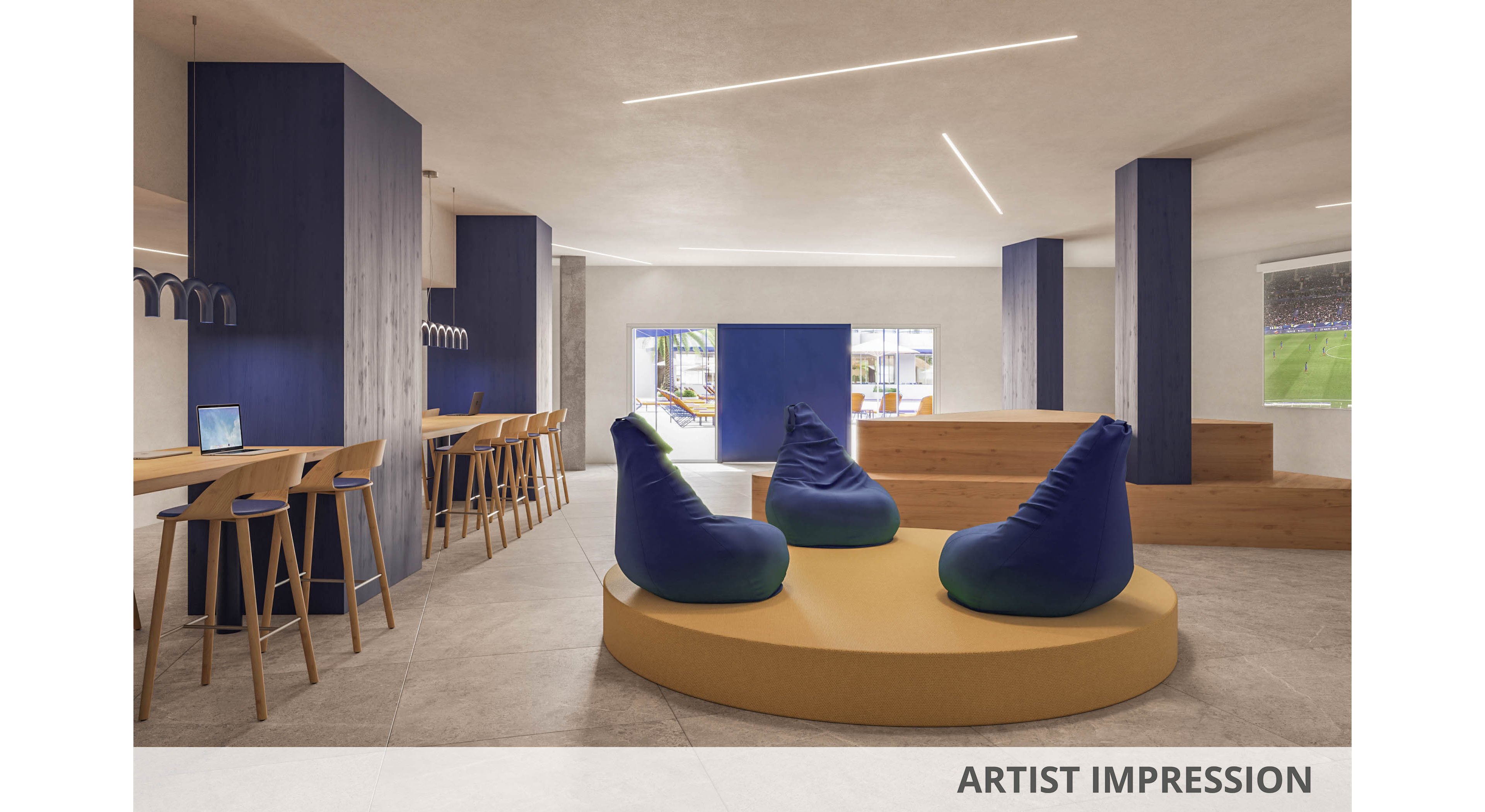 A contemporary open-plan lounge features a central circular platform with several bean bag chairs. Wooden bar-height workstations with stools line the left side, each equipped with laptops. Tall blue columns and minimalist lighting create a clean, modern aesthetic. Large windows and a screen showing a sports field add a relaxed, social atmosphere to the space.
