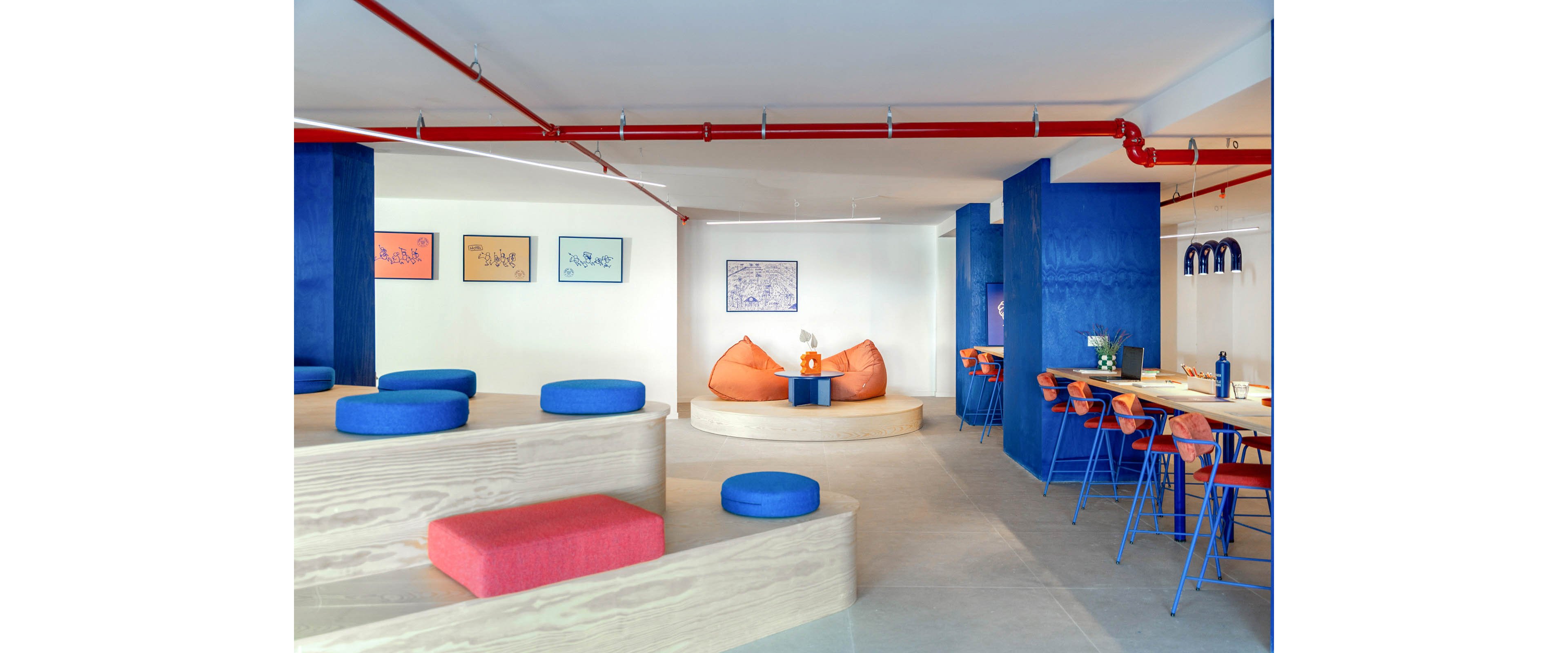 Bright contemporary lounge space features tiered wooden seating with colorful cushions and bean bags. A long shared desk with orange chairs lines the right side, creating an informal coworking area. Bold blue wall panels, red ceiling pipes, and framed artwork add graphic visual interest. The overall setting feels open, playful, and creative, suitable for casual meetings or relaxed work.