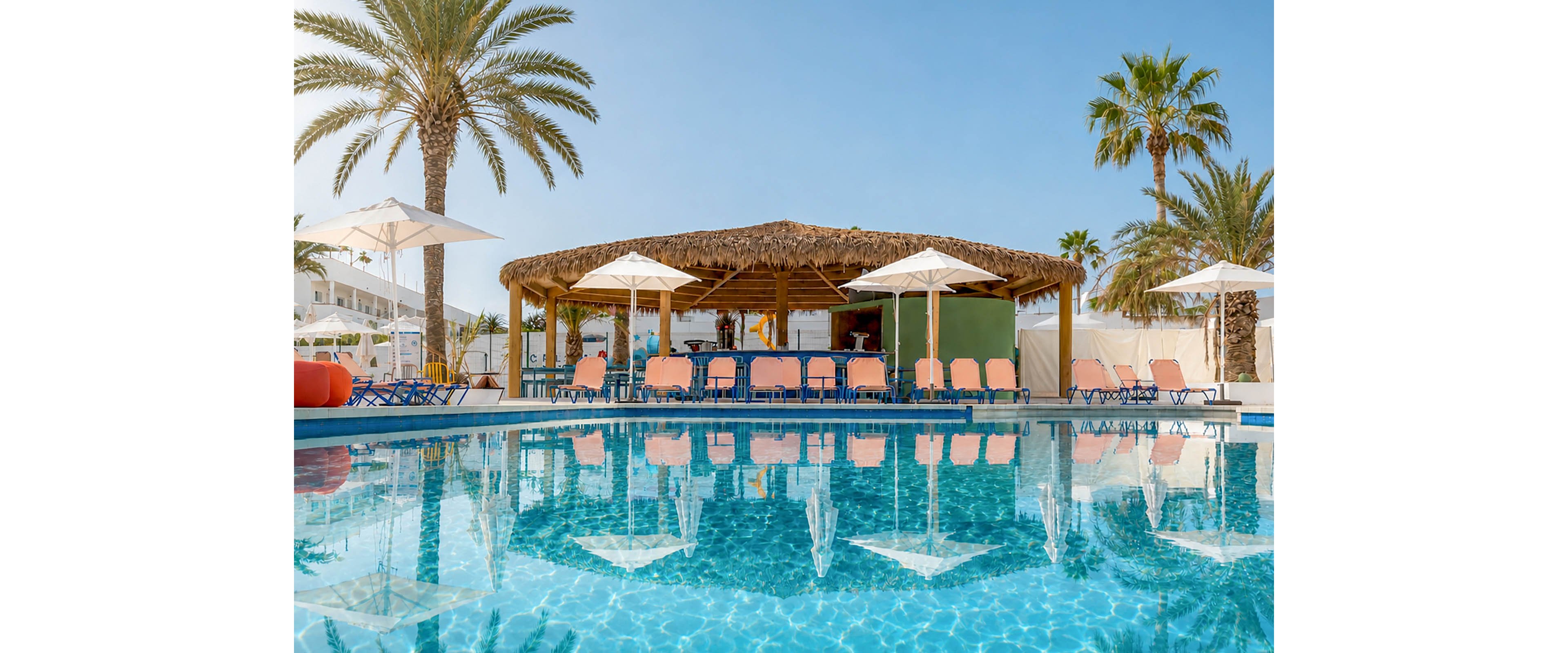 A clear blue swimming pool reflects palm trees and a thatched-roof bar in a sunny resort setting. Lounge chairs and umbrellas surround the pool, suggesting a relaxing vacation atmosphere. The sky is bright and cloudless, enhancing the warm, tropical feel. No people are visible, emphasizing the tranquil and inviting environment.