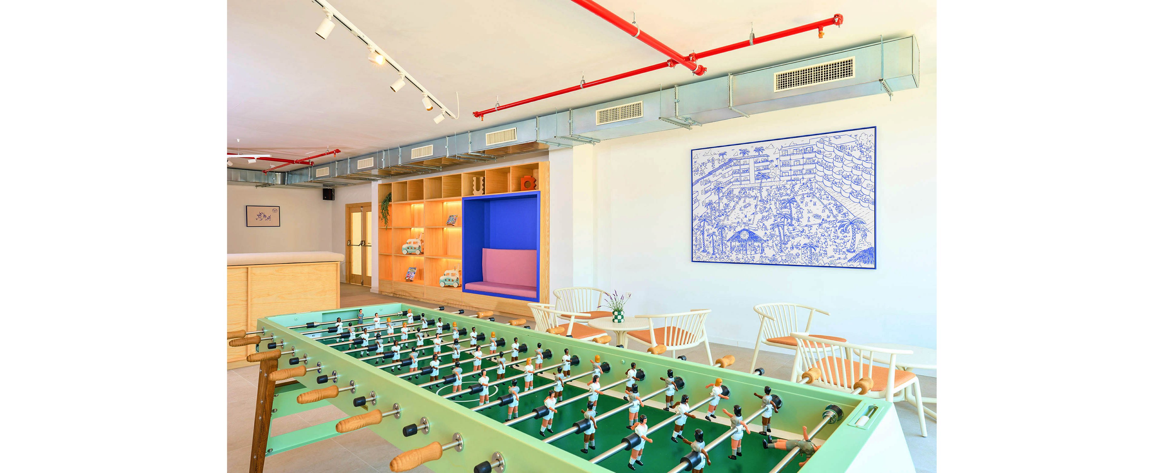 A large green foosball table dominates a bright, modern lounge area. The room features light wooden furniture, white chairs, and built-in shelving with decorative items. Overhead, exposed ductwork and red pipes add an industrial touch to the clean interior. A blue wall illustration and soft pastel accents create a playful, relaxed atmosphere.