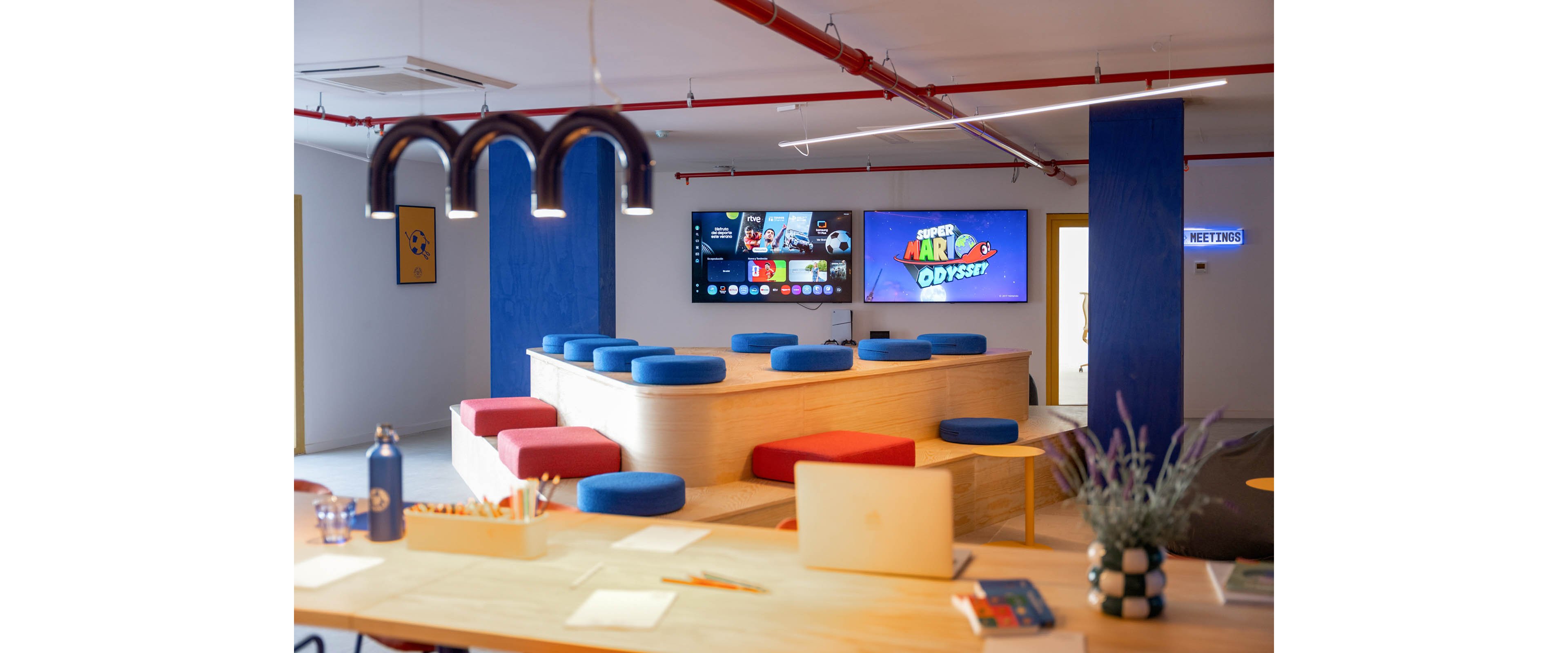 A contemporary lounge space features tiered wooden seating with blue and red cushions. Multiple wall-mounted screens display a media interface and a game titled "Super Mario Odyssey." A long table in the foreground holds a laptop, water bottle, pencils, and decorative plant under stylish pendant lights. Bright blue columns, yellow doors, and playful decor create a casual, creative atmosphere, with a sign reading "MEETINGS" visible on one door.