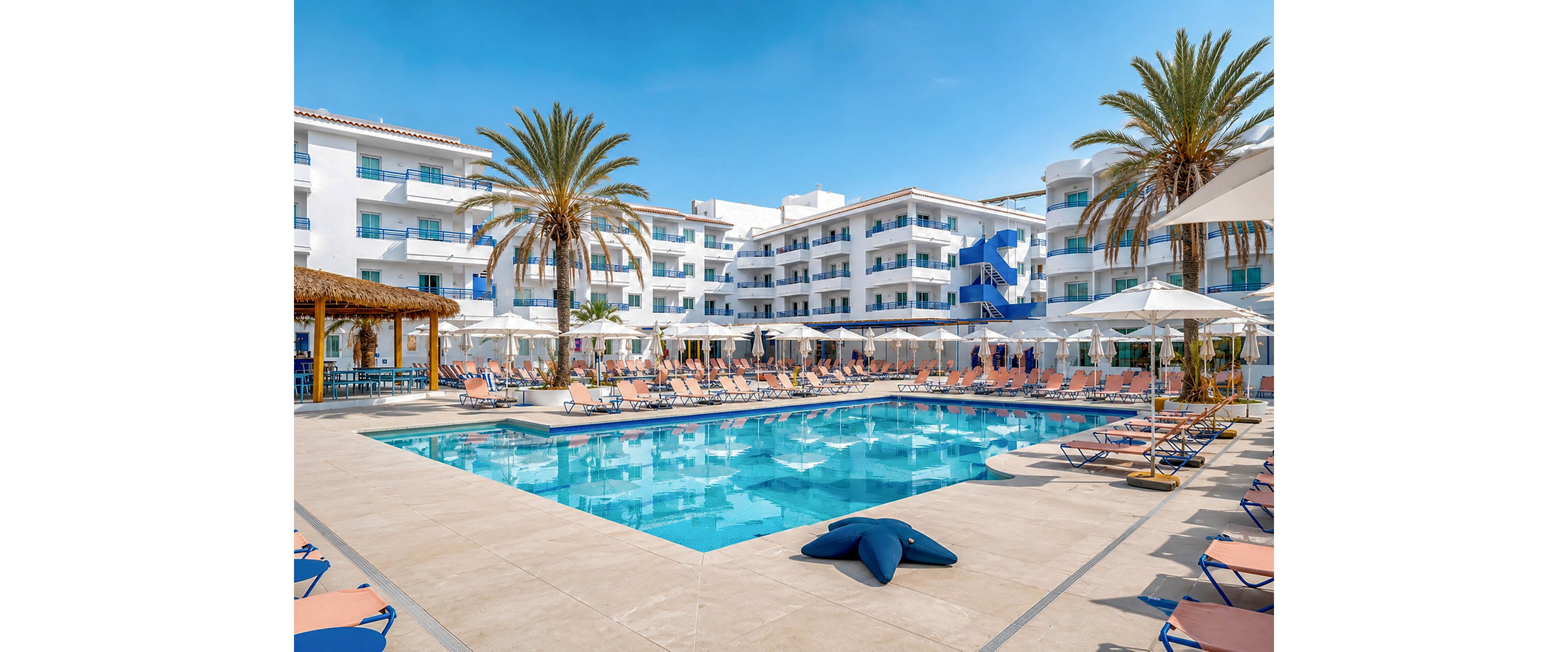 A large outdoor swimming pool sits at the center of a modern resort hotel courtyard. Rows of sun loungers and white umbrellas surround the pool, creating a relaxed vacation atmosphere. Tall palm trees rise above the pool area, and multi-story white buildings with balconies frame the scene. The clear blue sky and reflections in the water emphasize a bright, sunny day at a coastal or island destination.