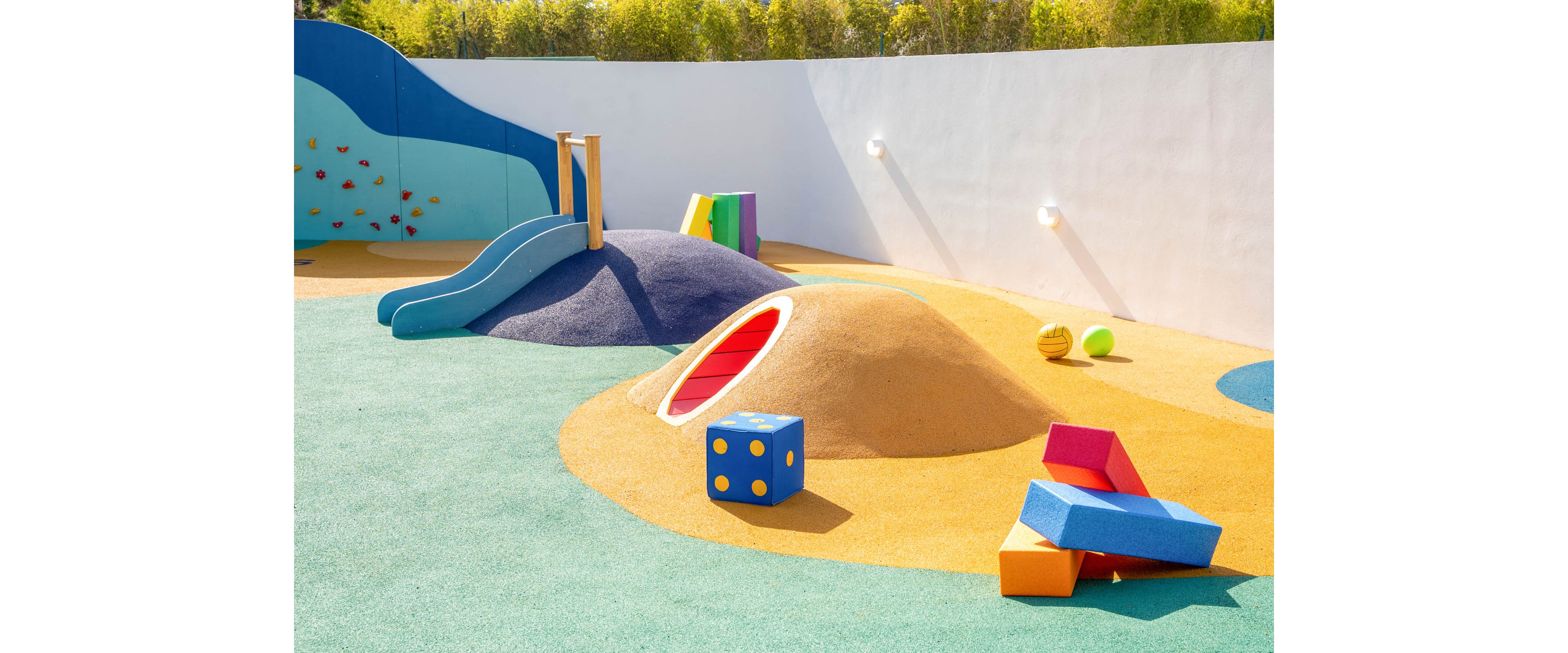 A bright, contemporary playground features sculpted mounds, a small slide, and tunnel openings set on cushioned flooring. The space is outdoors, enclosed by a white wall and backed by greenery, under clear sunny light. Large geometric play blocks and balls in vivid primary colors add visual interest and invite imaginative play. The overall design emphasizes safety, creativity, and a cheerful atmosphere for children.