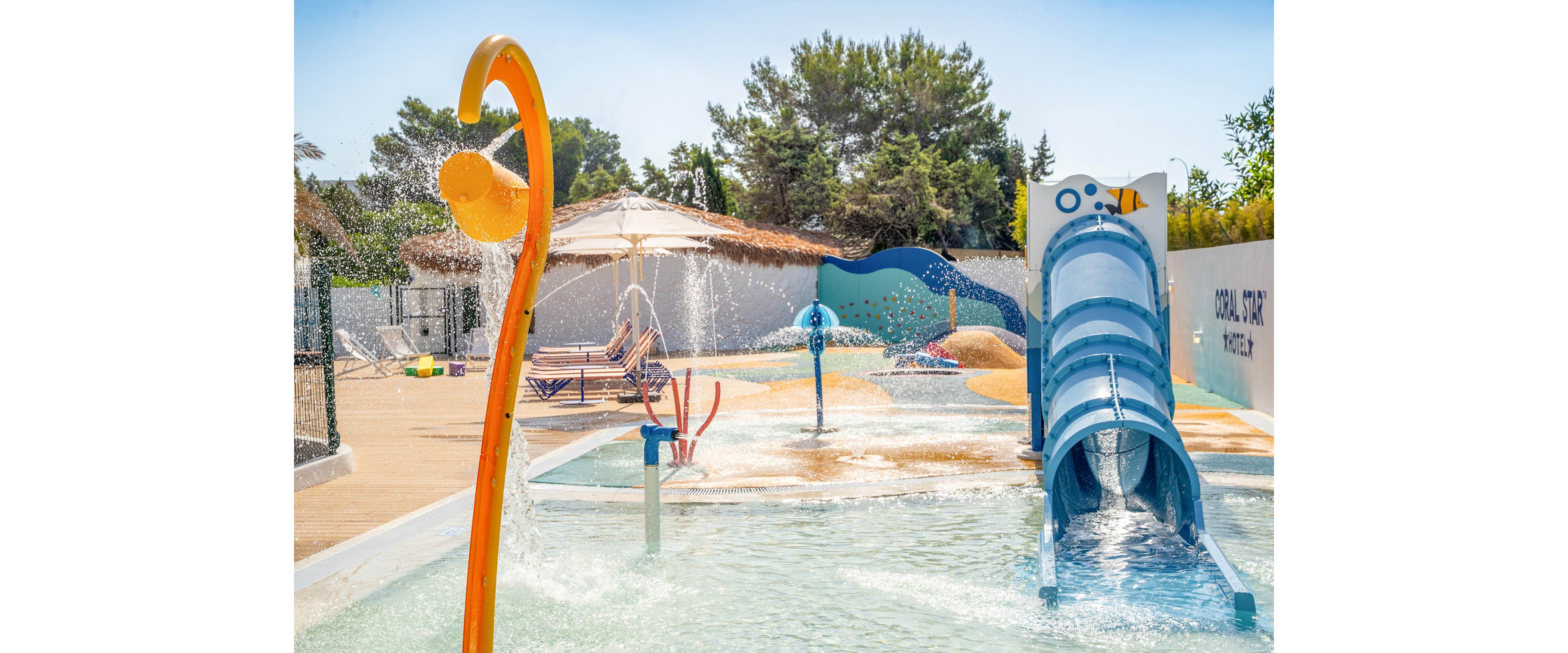 An outdoor splash park features a bright blue tube water slide and multiple spray fountains. Lounge chairs and umbrellas sit nearby on the poolside deck, suggesting a family-friendly recreation area. Trees and a clear blue sky in the background create a sunny, vacation-like atmosphere. The scene emphasizes playful water features, vibrant colors, and a relaxed summer mood.