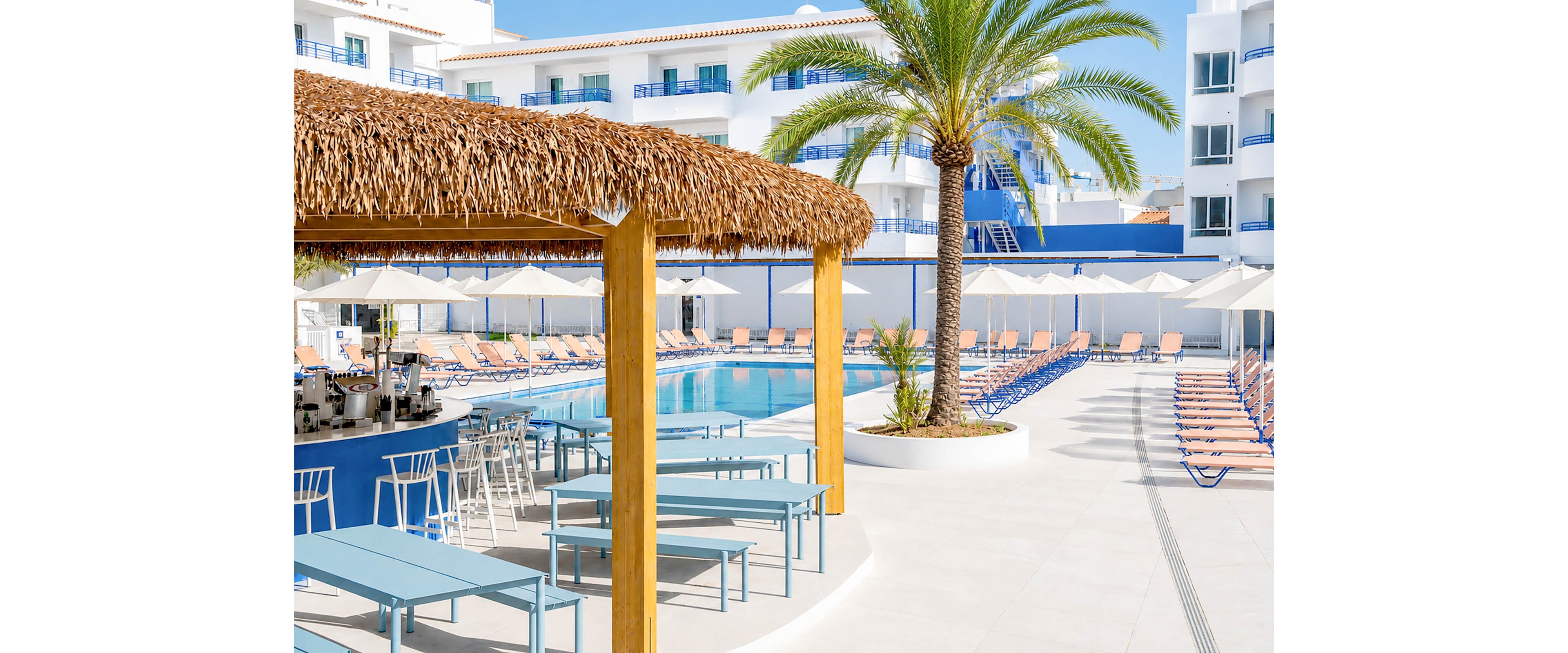 An outdoor resort swimming pool is surrounded by rows of orange lounge chairs and white umbrellas. A thatched-roof poolside bar with wooden posts and blue tables sits in the foreground. A tall palm tree rises beside the bar, adding a tropical feel to the bright, sunny courtyard. White multi-story buildings with blue railings frame the scene, creating a clean and modern holiday atmosphere.
