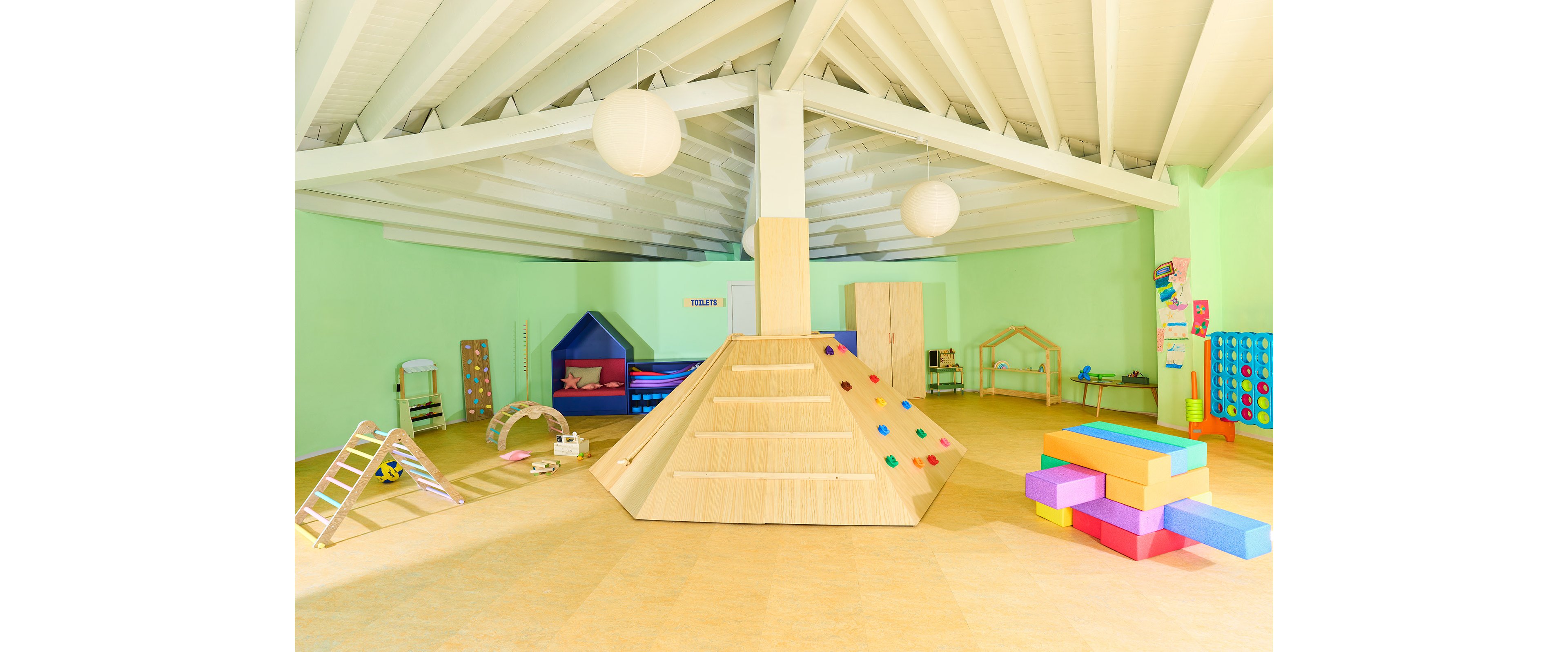 Colorful indoor playroom features a large wooden climbing pyramid at the center. Soft play blocks, slides, and arches are arranged around the room, creating a child-friendly activity space. The walls are painted light green, with pastel play equipment and geometric structures adding visual interest. Round ceiling lights and exposed beams give the space a modern, airy feel.