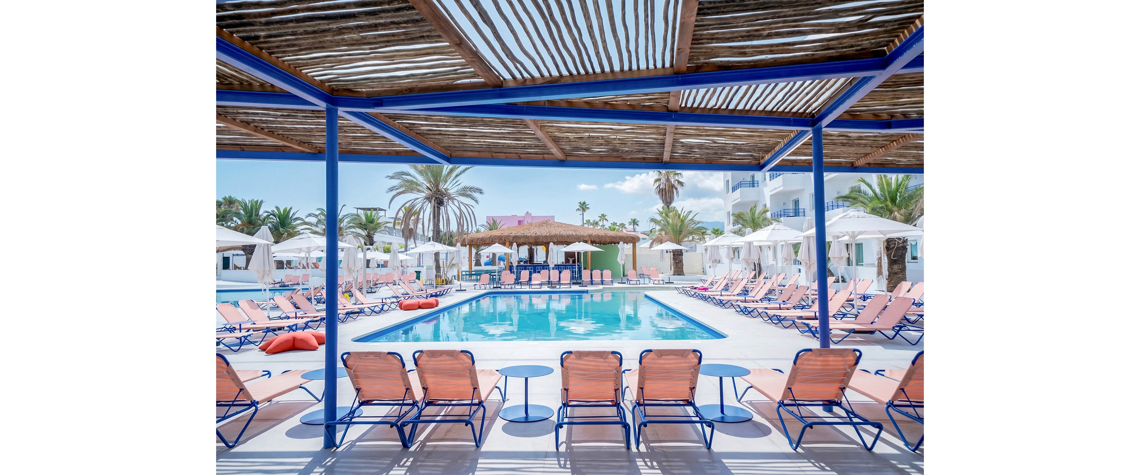 An inviting outdoor swimming pool is surrounded by rows of pastel-colored lounge chairs under a shaded pergola. White cabanas and umbrellas line the poolside area, creating a relaxed resort atmosphere. Palm trees and bright blue skies frame the scene, with a central bar hut visible in the background. The overall setting feels summery, vibrant, and ideal for vacation leisure.