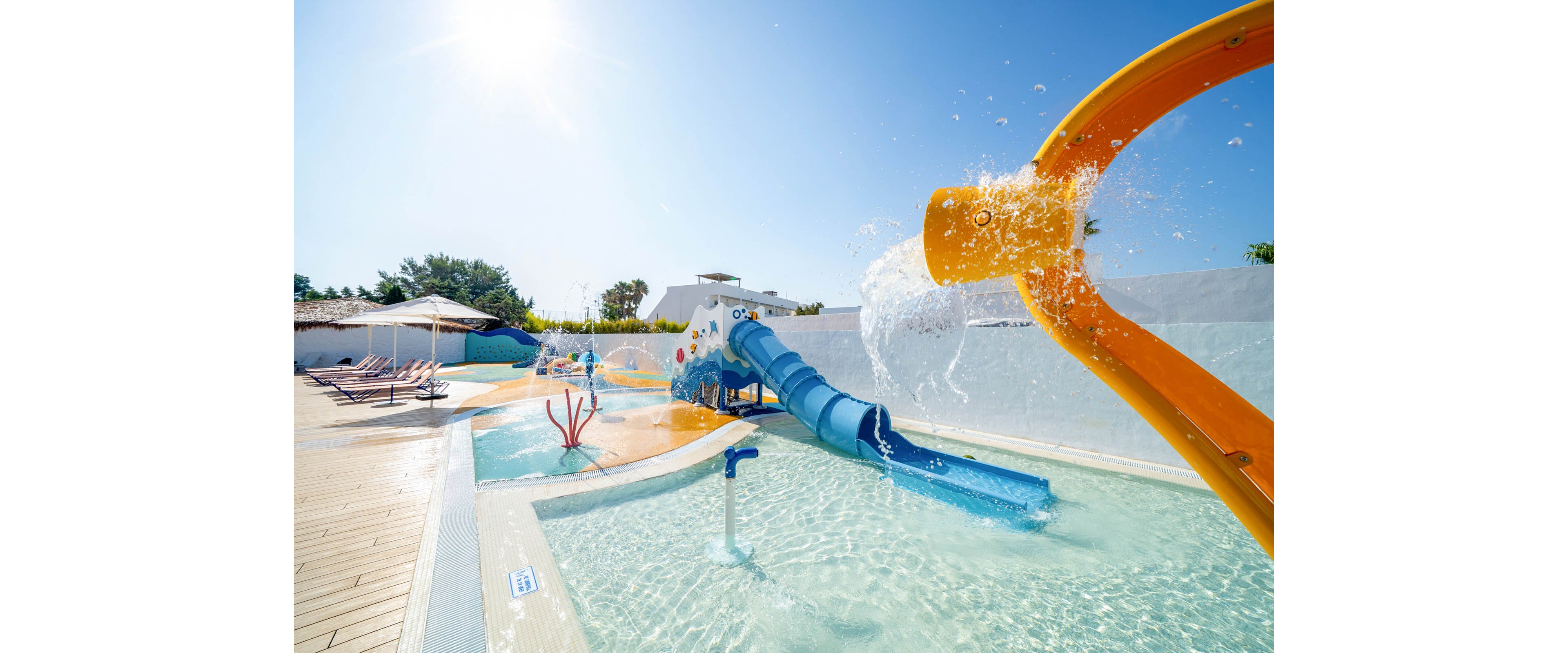An outdoor water play area features a shallow pool with colorful slides and splash structures under a clear blue sky. A large yellow water spout pours water into the pool, while a winding blue slide extends from a small platform. Lounge chairs and umbrellas line the deck in the background, suggesting a relaxing family-friendly environment. The bright colors and sunlight create a cheerful, summery atmosphere.