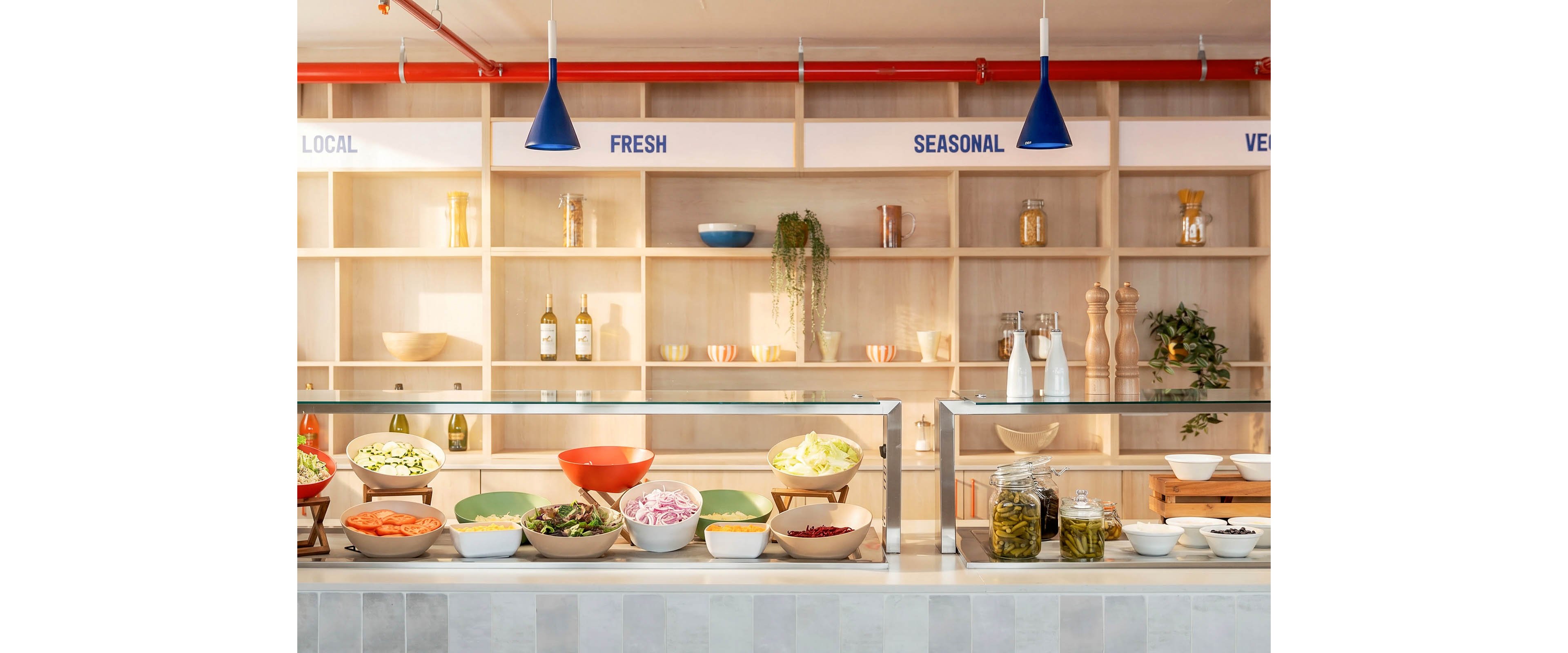 A bright, modern salad bar displays a variety of fresh vegetables and toppings in bowls along a glass counter. Behind the counter, open wooden shelves hold bottles, jars, plants, and serving accessories, creating a clean, organized backdrop. Blue pendant lights hang from the ceiling, and the words "LOCAL", "FRESH", and "SEASONAL" are clearly visible on the upper wall. The overall scene suggests a contemporary, health-focused dining environment with an emphasis on natural ingredients.