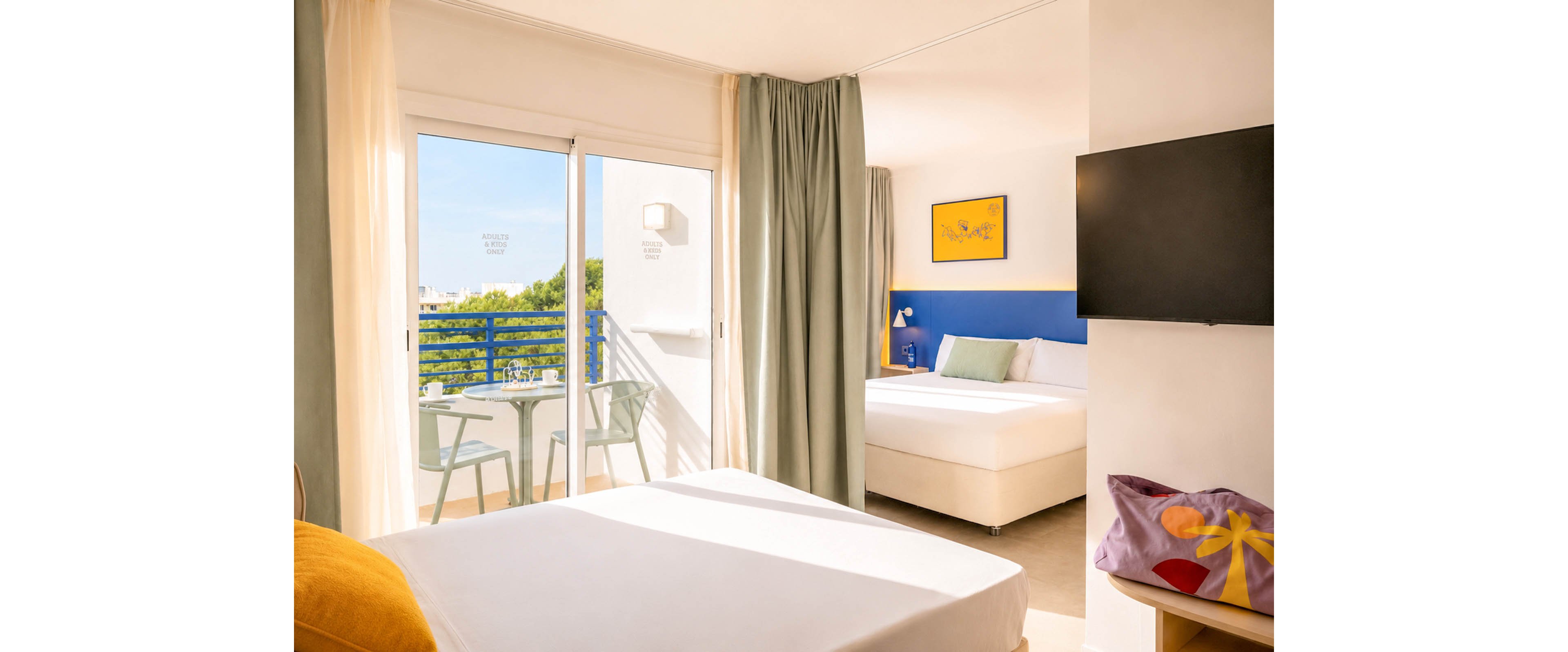 Sunlit hotel room features two white beds separated by a curtain, creating a cozy and flexible sleeping area. Large sliding glass doors open to a small balcony with two chairs and a table, overlooking an outdoor view with greenery and blue railings. The interior decor includes a blue accent wall, soft neutral curtains, and a wall-mounted television, giving the space a modern and inviting feel. Warm daylight enhances the clean, minimalist style and relaxed vacation atmosphere.