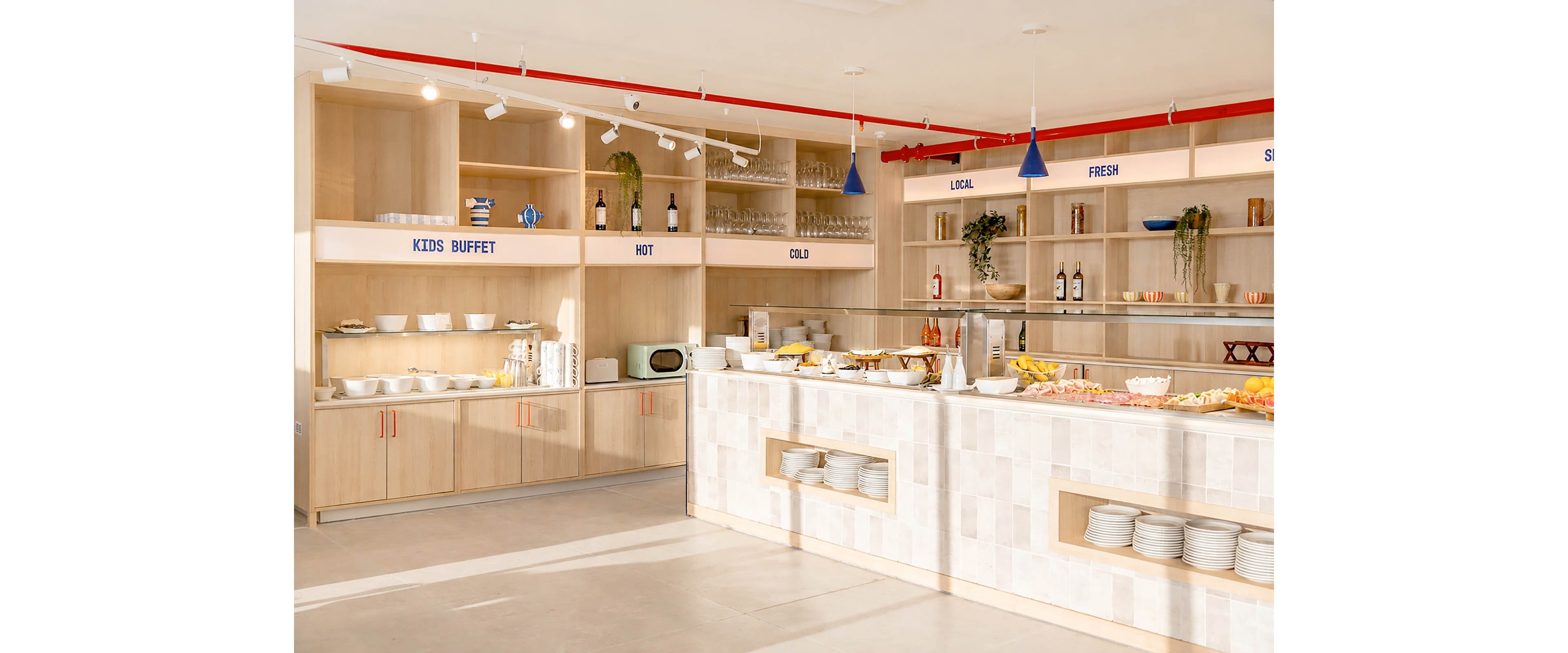 A modern self-service buffet counter is set up in a bright, minimalist restaurant interior. Wooden shelves and cabinets display plates, bowls, glasses, and bottles, with food trays arranged along the main counter. Blue signage labels different sections including KIDS BUFFET, HOT, COLD, LOCAL, and FRESH. The space is lit by ceiling spotlights and blue pendant lamps, creating a clean and welcoming atmosphere.