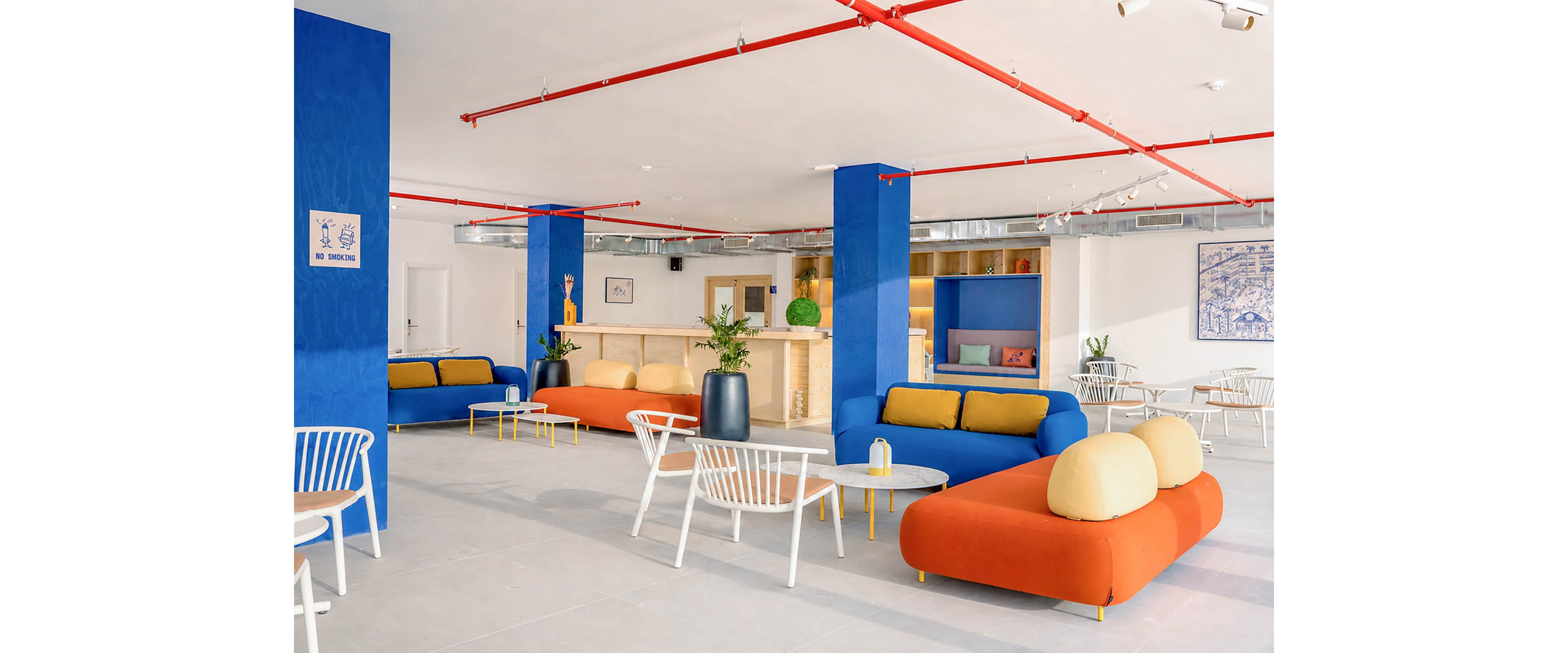 Bright contemporary lounge area featuring vivid blue columns and low orange sofas with yellow cushions. White chairs and round tables are arranged across the open space, creating a casual seating layout. A light wood bar counter with decorative plants and shelves sits at the back of the room. Red ceiling pipes and minimalist artwork add graphic accents to the clean, modern interior.