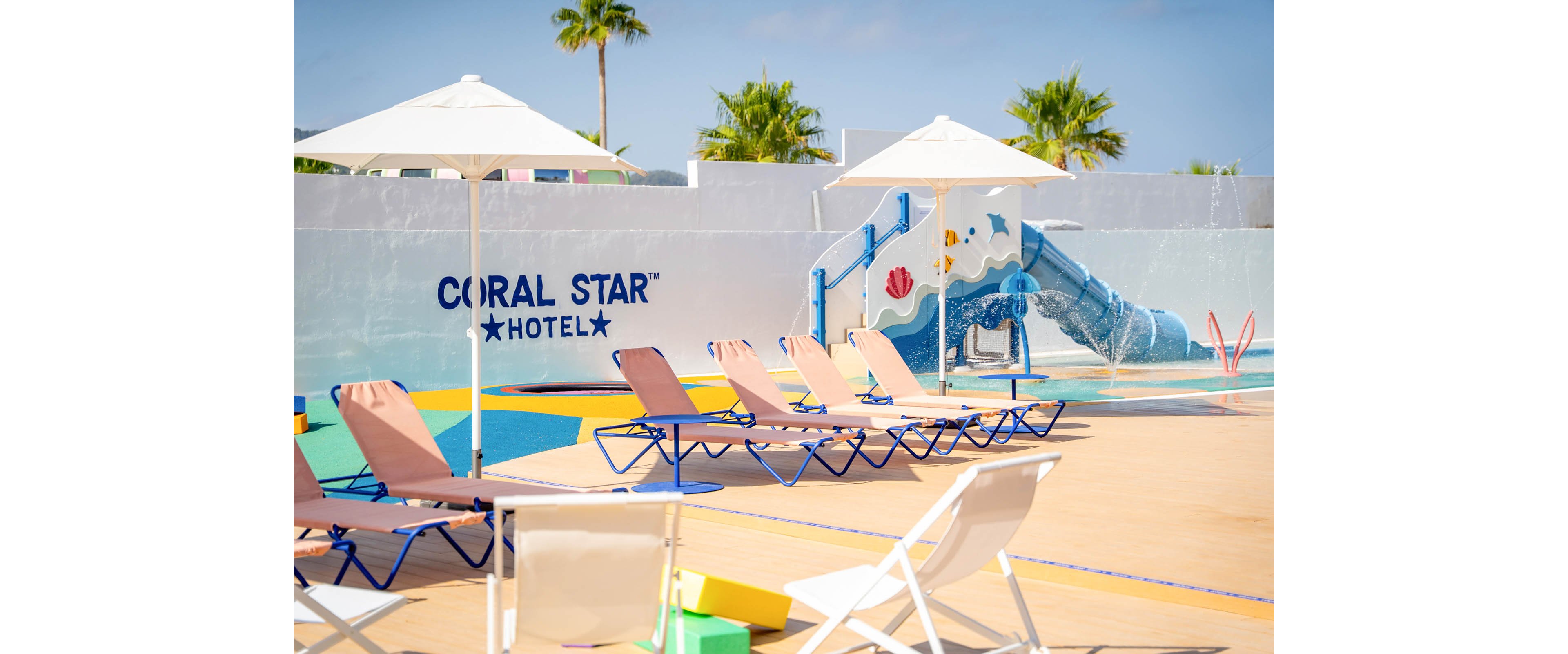 Outdoor hotel pool area with multiple empty sun loungers arranged under white parasols. A colorful children’s water playground with a blue slide and splash features stands in the background. The words “CORAL STAR HOTEL” are clearly visible on the wall beside playful sea-themed graphics. Palm trees and a bright blue sky create a warm, relaxing vacation atmosphere.