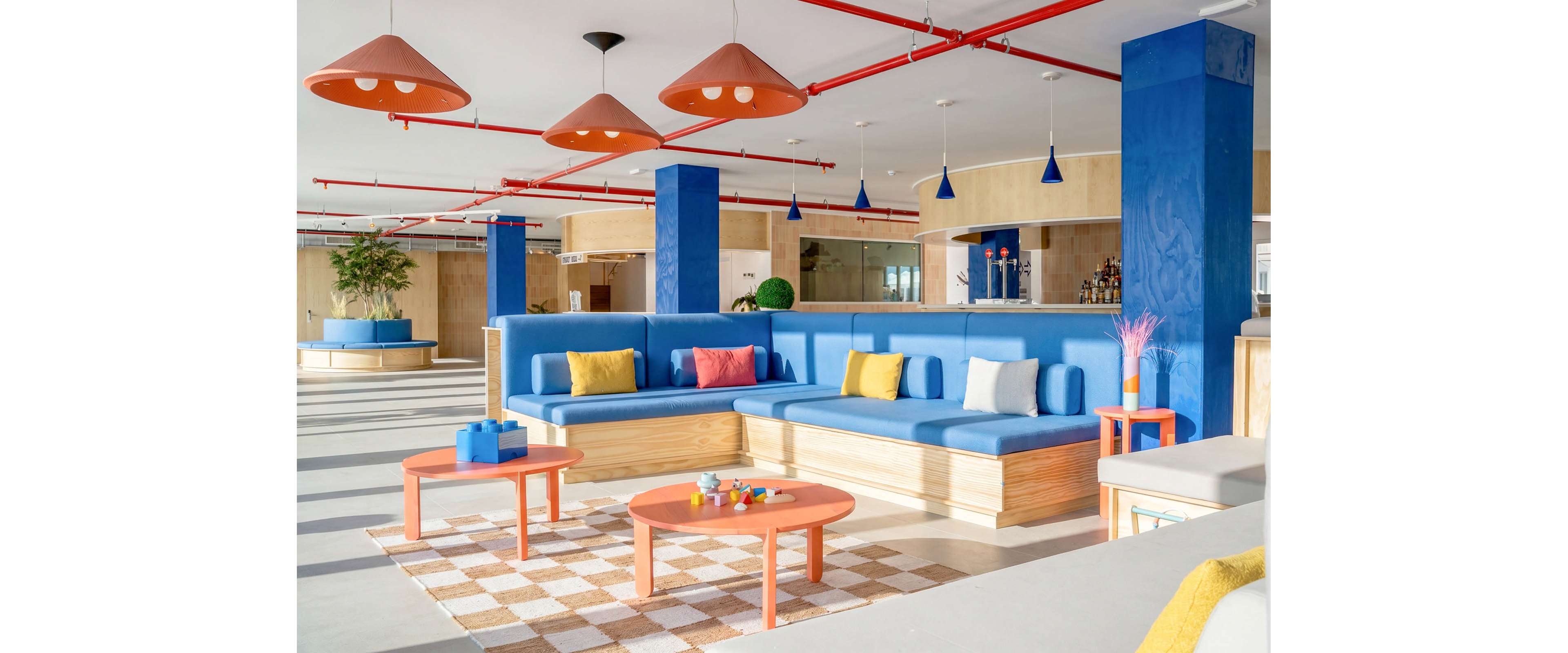 A bright contemporary lounge features large blue upholstered bench seating with wooden bases and colorful accent pillows. Coral coffee tables and matching pendant lights add playful contrast to the open interior. Blue columns, red ceiling pipes, and a checkered rug create a bold geometric look. Natural light floods the space, highlighting the casual, social atmosphere and vibrant decor.