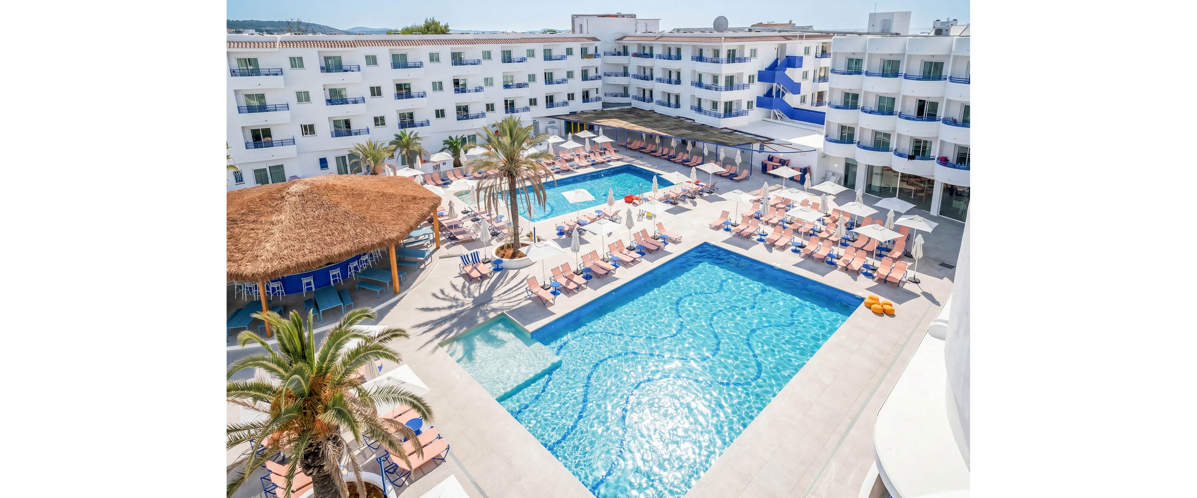 A large hotel resort courtyard features several bright blue swimming pools surrounded by sun loungers and umbrellas. White multi-story buildings with balconies frame the open space, creating a modern vacation setting. A thatched-roof bar area and palm trees add a tropical atmosphere to the scene. The overall mood is sunny, relaxed, and inviting for holiday travelers.