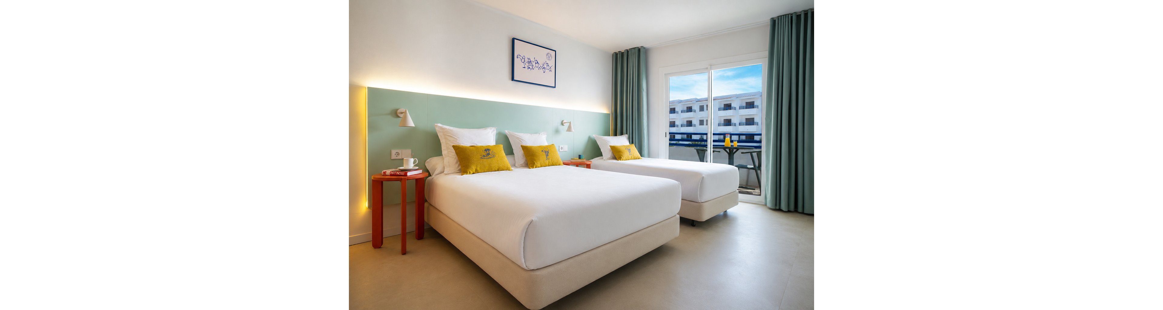 Contemporary hotel room featuring two neatly made twin beds with white linens and yellow accent pillows. The space is decorated in soft pastel tones with a mint green accent wall, minimalist bedside lamps, and small red side tables. Large windows with teal curtains open to a balcony overlooking a white building complex under a blue sky. Overall, the room feels clean, airy, and inviting, ideal for vacation or business stays.