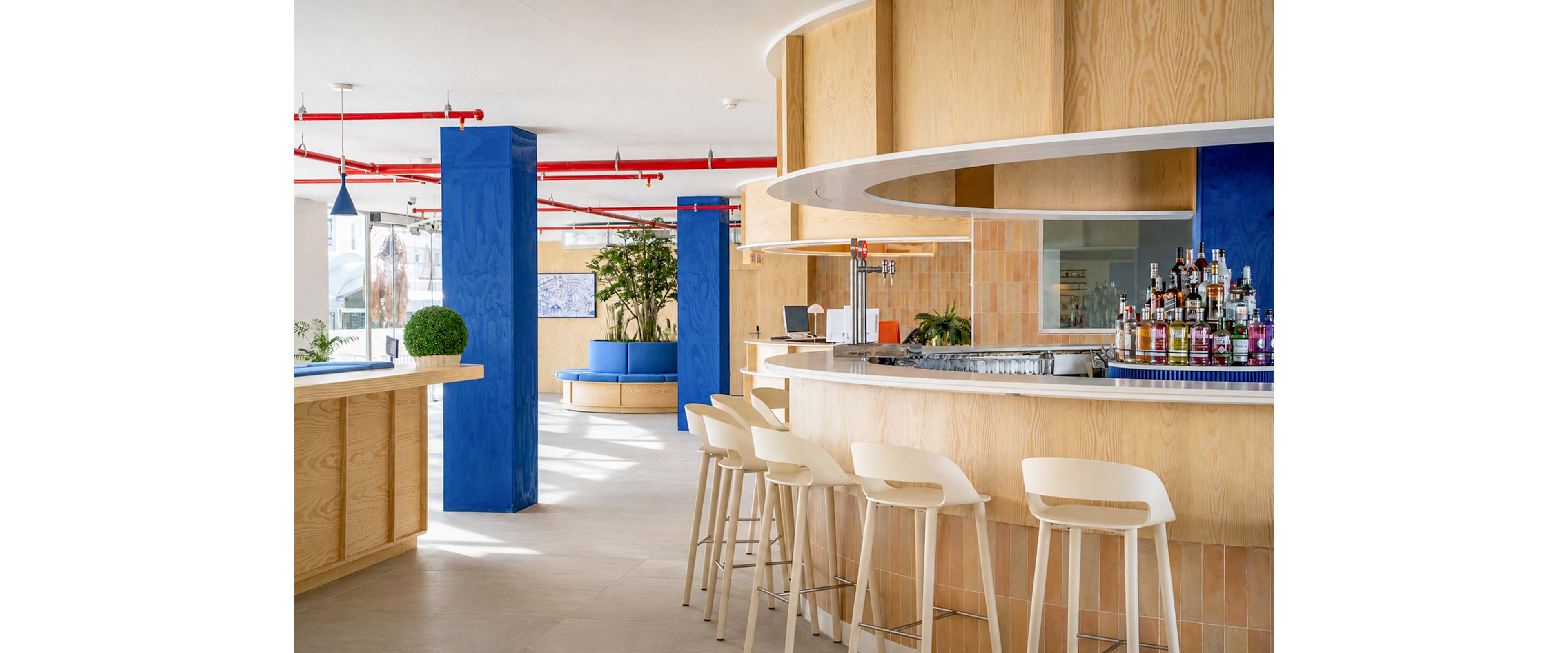 Contemporary bar interior features a curved counter with white stools and a display of liquor bottles. Bright blue columns and trim contrast with light wood surfaces, creating a fresh, coastal atmosphere. Large windows and indoor plants add natural light and greenery to the open space. The setting appears clean, minimal, and inviting for socializing or relaxing.