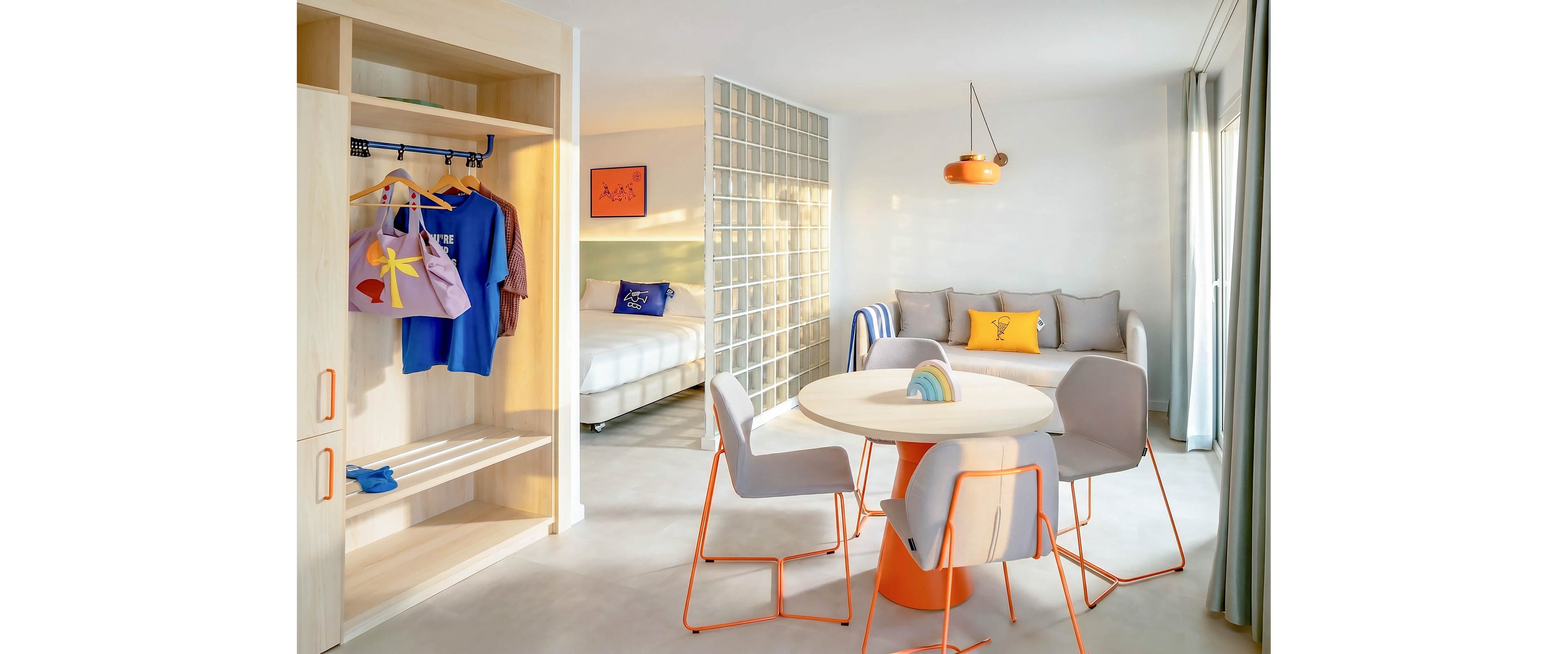 A bright modern hotel suite features an open-plan layout with a small dining table and pastel-colored chairs in the foreground. To the left, an open wardrobe displays hanging clothes and bags, while a cozy bed is partially visible behind a glass block partition. The room is decorated with playful cushions, wall art, and an orange pendant light that complement the soft neutral walls and floor. Large windows with sheer curtains allow natural light to fill the contemporary interior.