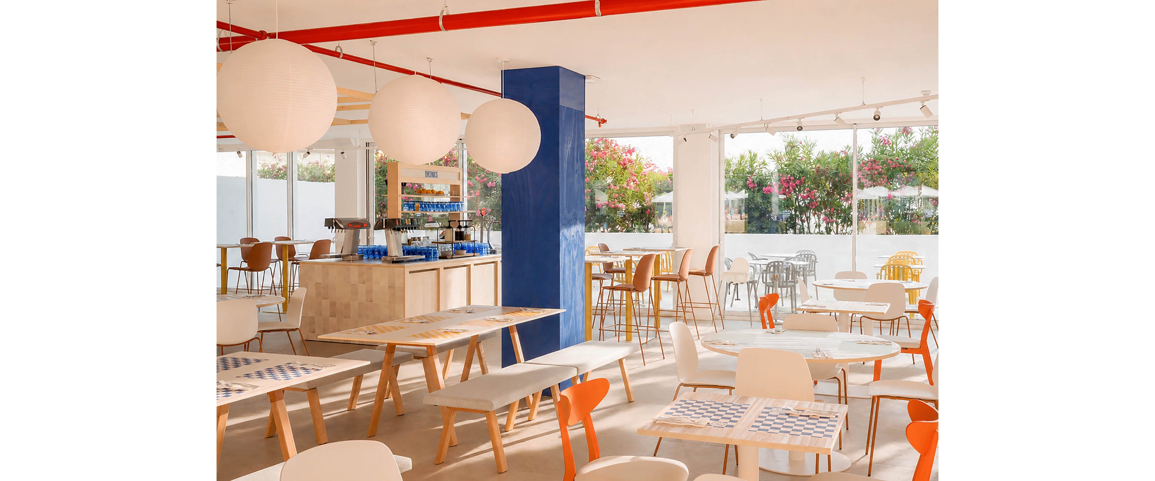 A contemporary café interior features light wooden tables and mixed chairs in white and orange tones. Large spherical pendant lights hang from the ceiling, and a bold blue column stands near the central counter area. Floor-to-ceiling windows reveal blooming plants and outdoor seating, filling the space with natural light. The overall atmosphere is airy, cheerful, and inviting for casual dining or coffee breaks.