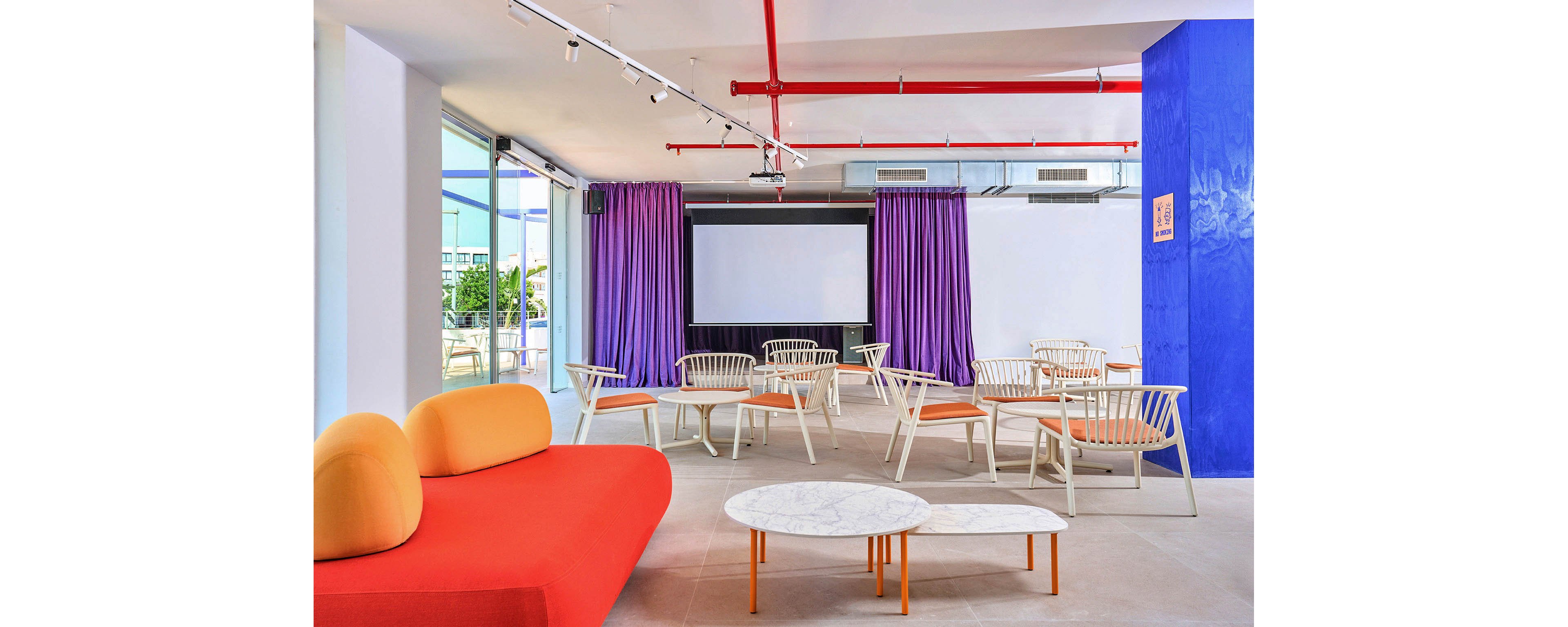 A bright contemporary lounge features a large projection screen at the front of the room. White chairs with orange cushions are arranged facing the screen, creating a casual presentation or meeting area. A bold orange sofa and round white marble coffee tables add playful contrast to the space. Purple curtains, a vivid blue accent wall, and exposed red ceiling pipes contribute to the colorful, modern aesthetic.
