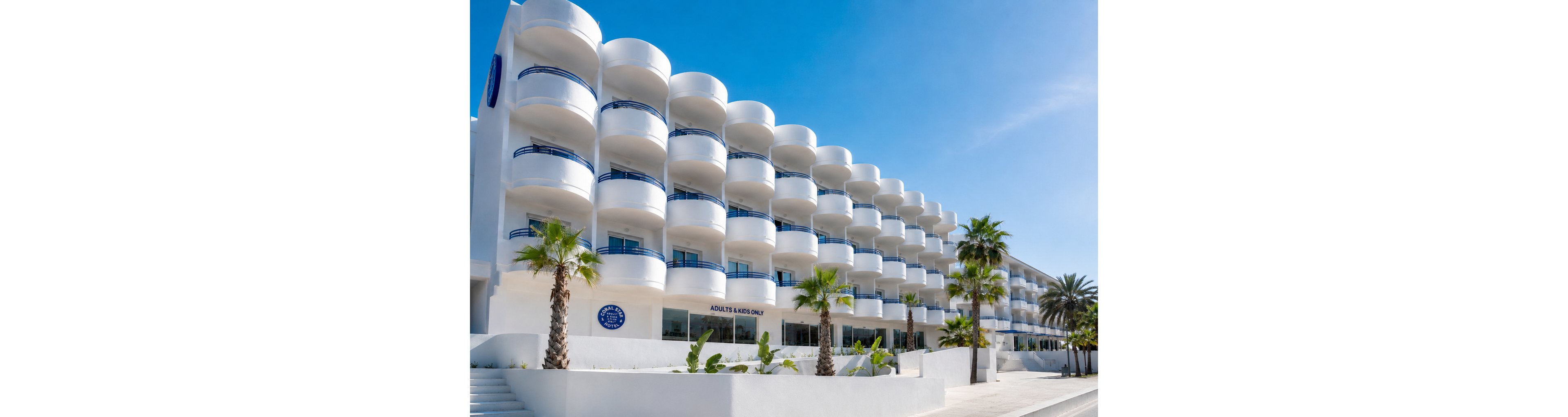 A contemporary white hotel building features multiple rounded balconies facing a sunny street. Palm trees and low tropical plants line the sidewalk, enhancing the coastal atmosphere. Blue railings and accents contrast with the bright white facade under a clear blue sky. Visible text on the building reads "ADULTS KIDS ONLY" near the entrance.