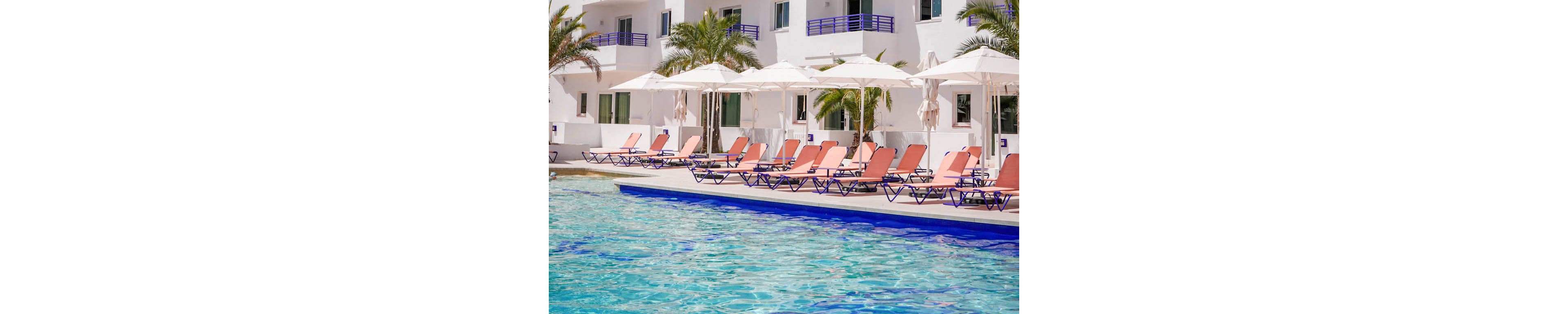 A contemporary white hotel building with multiple balconies overlooks a clear blue swimming pool. Rows of sun loungers and white umbrellas line the poolside terrace, suggesting a relaxing vacation setting. Palm trees add a tropical touch against the bright blue sky. An orange flower-shaped seat in the foreground adds a playful pop of color to the scene.