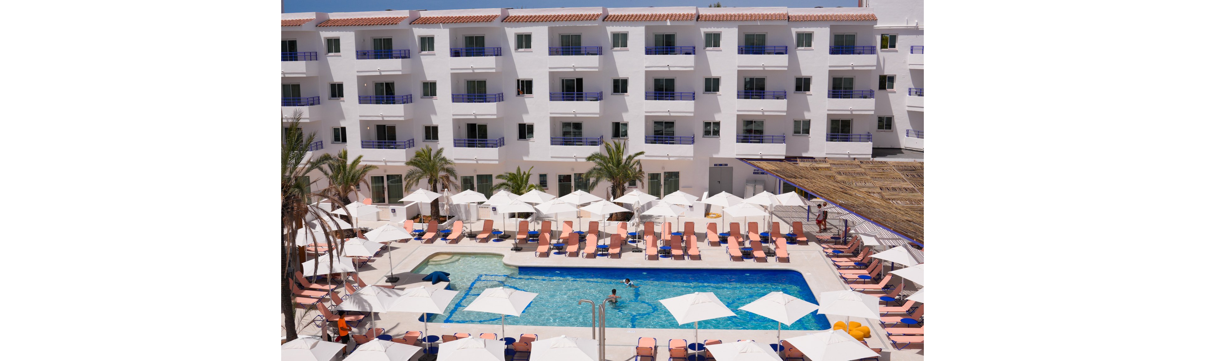A large rectangular outdoor swimming pool sits at the center of a modern white hotel courtyard. Rows of pink lounge chairs and white umbrellas surround the pool, creating a resort-style relaxation area. A few guests enjoy the water while others rest on the sunbeds. Palm trees and multiple balconies add to the vacation atmosphere under bright daylight.