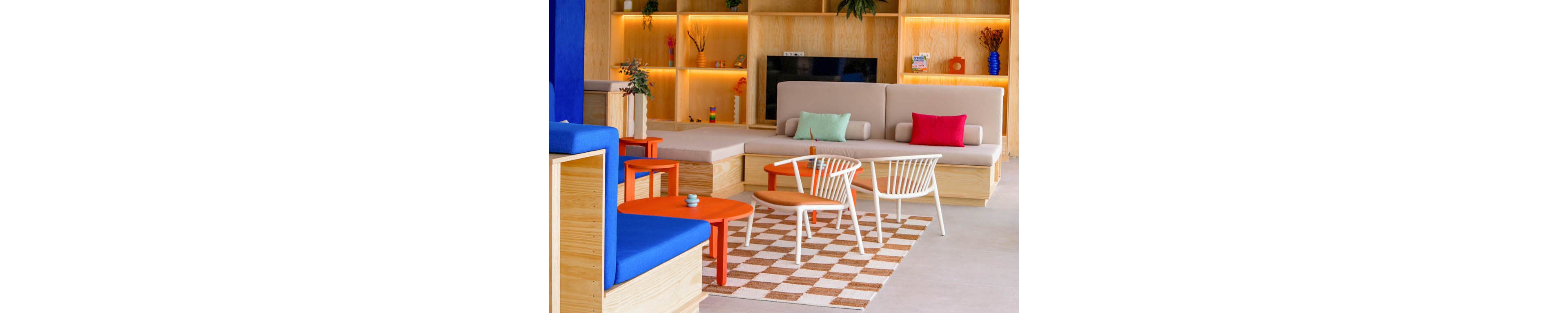A contemporary lounge space features built-in wooden seating and bright accent cushions. The room includes a checkered floor section with a small table and white chairs in the center. Warm backlit shelves display decorative objects and plants along the wall. Bold red ceiling lights and exposed pipes add an industrial touch to the clean, modern interior.