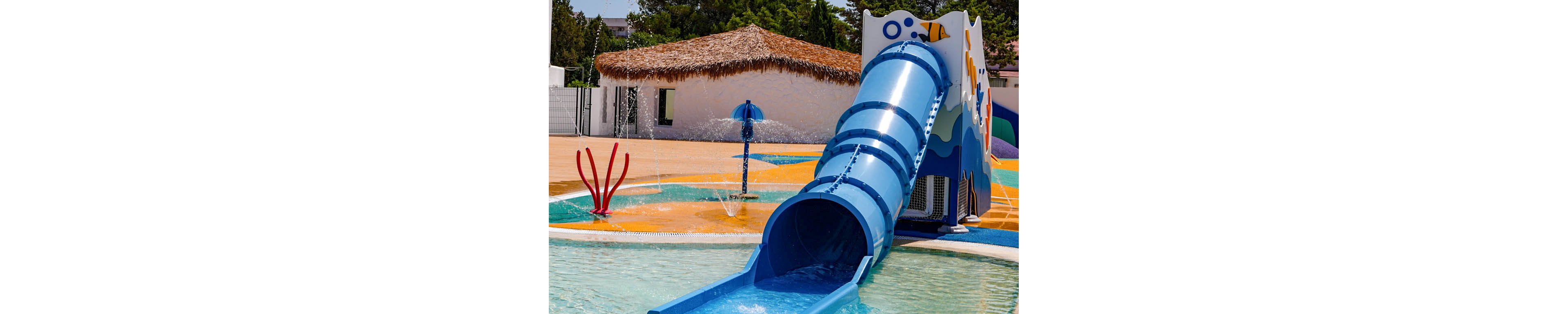 A bright blue tube waterslide ends in a shallow pool at an outdoor splash park. The slide structure is decorated with playful shapes and surrounded by interactive water features. Clear water flows from the slide into the pool under a sunny blue sky. Trees and a thatched-roof building are visible in the background, suggesting a warm resort or recreational setting.
