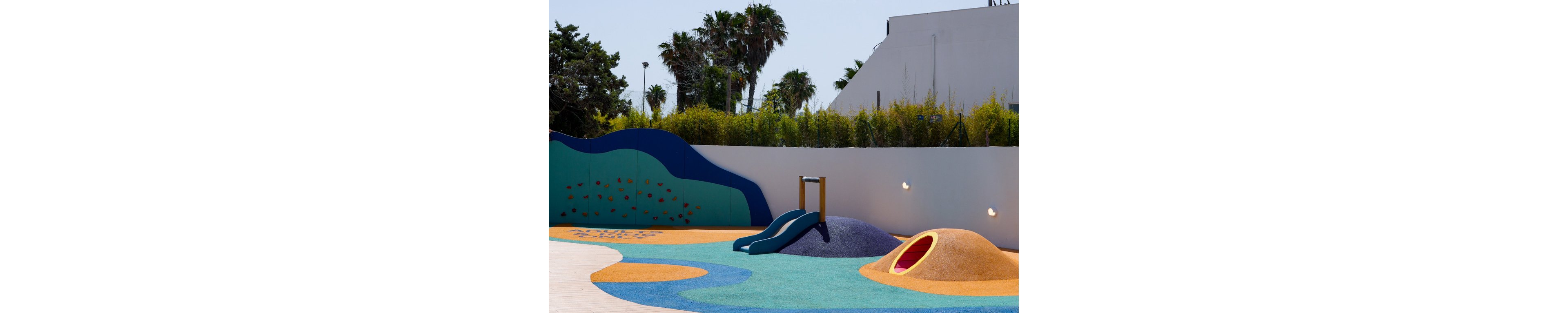 A modern outdoor playground features a small slide and sculpted play mounds in bright colors. The play area has a smooth wooden deck and soft, rubberized flooring in blue, yellow, and green tones. In the background, white contemporary buildings and tall palm trees stand against a clear blue sky. The scene is sunny and calm, with no people present.