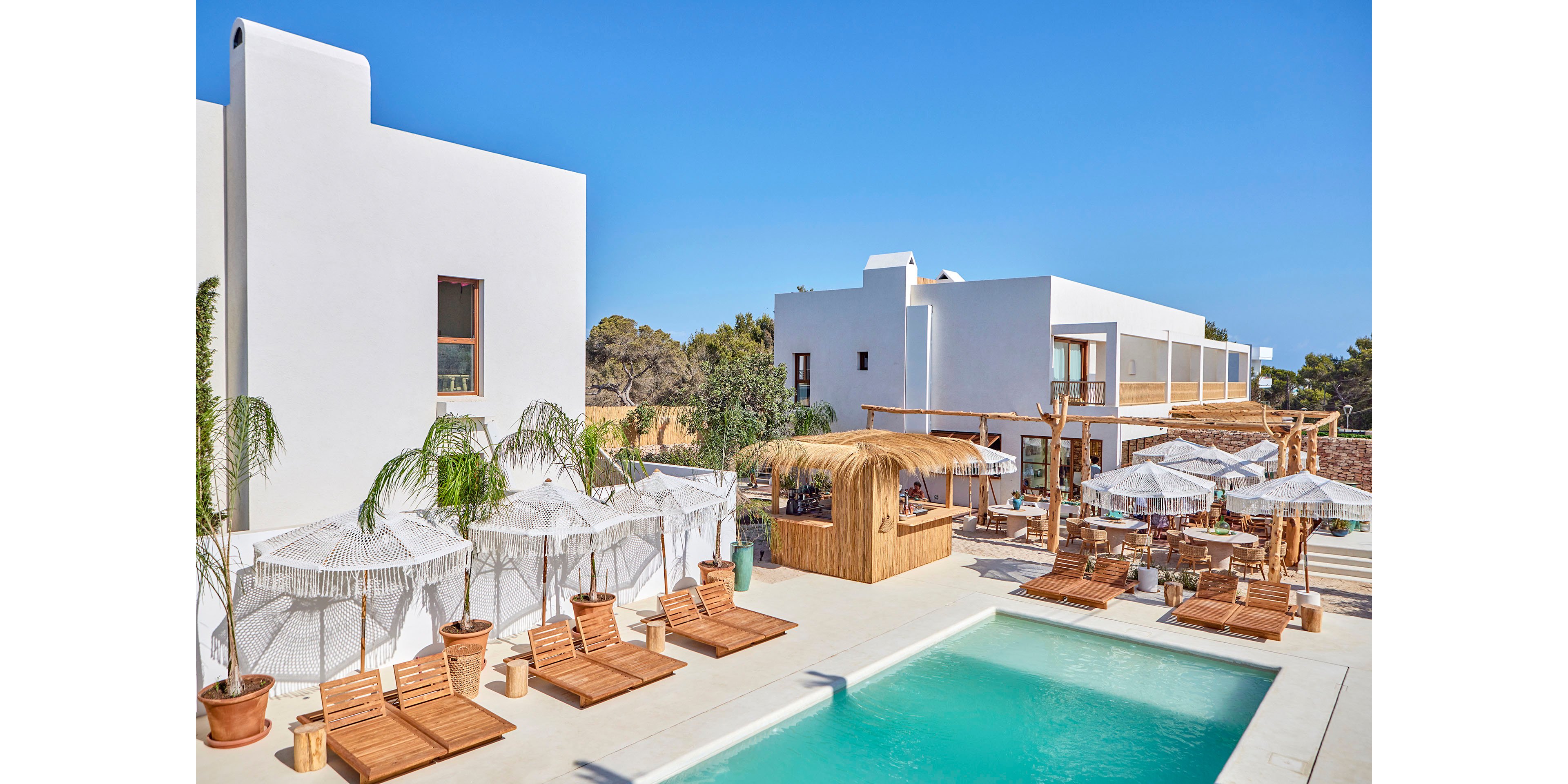 A contemporary white-walled resort features a turquoise swimming pool surrounded by wooden loungers and shaded cabanas. A thatched-roof bar and outdoor dining area create a relaxed vacation atmosphere. Palm trees and potted plants add greenery to the bright, sunlit courtyard. The clear blue sky and minimalist architecture emphasize a clean, modern Mediterranean style.