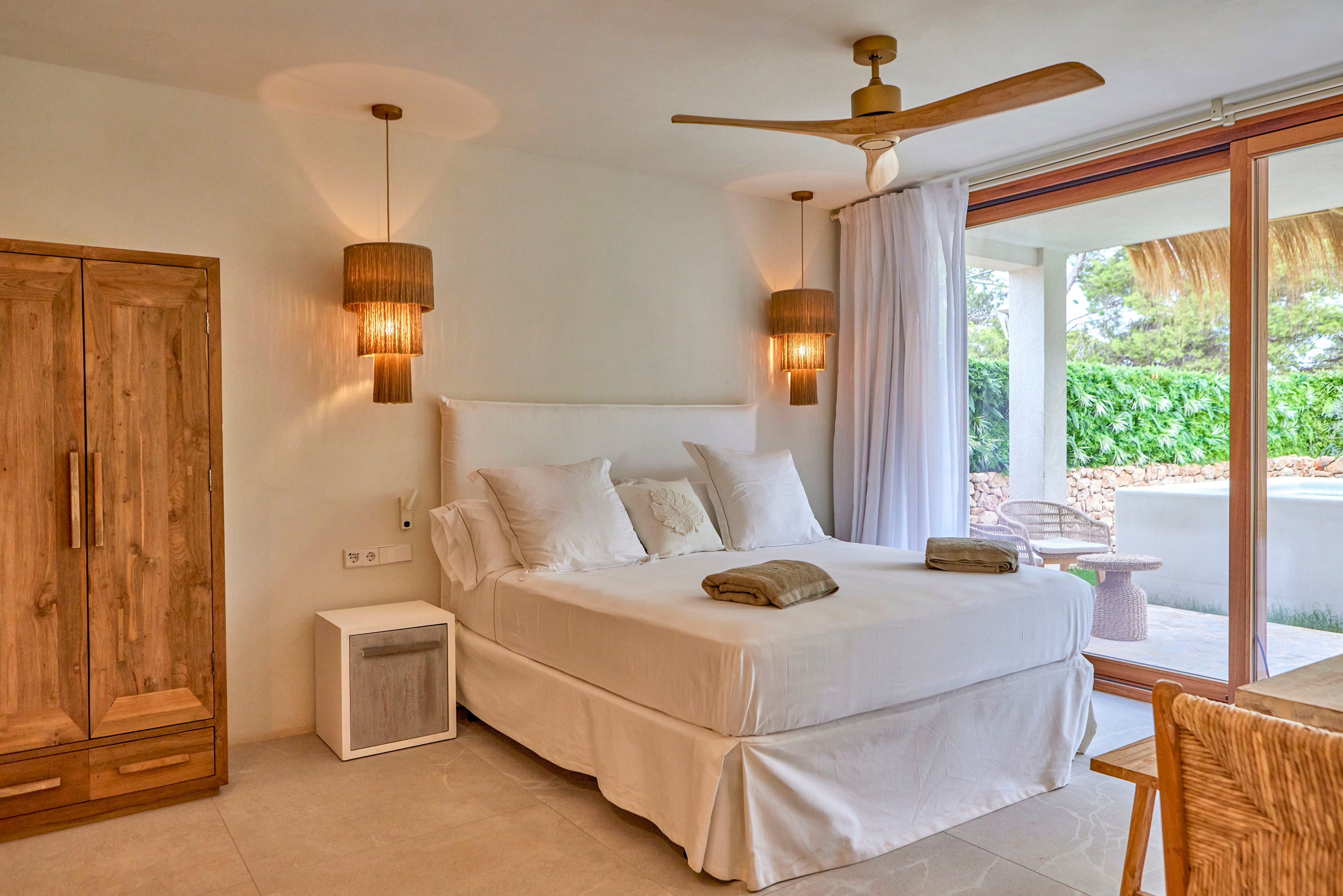 A bright, minimalist bedroom features a neatly made double bed with white linens and pillows. Warm wooden elements include a wardrobe, bedside table, and woven chair, complemented by matching pendant lights. Large glass doors open to a lush green outdoor area, allowing natural light to fill the room. Neutral tones and simple decor create a calm, contemporary atmosphere.