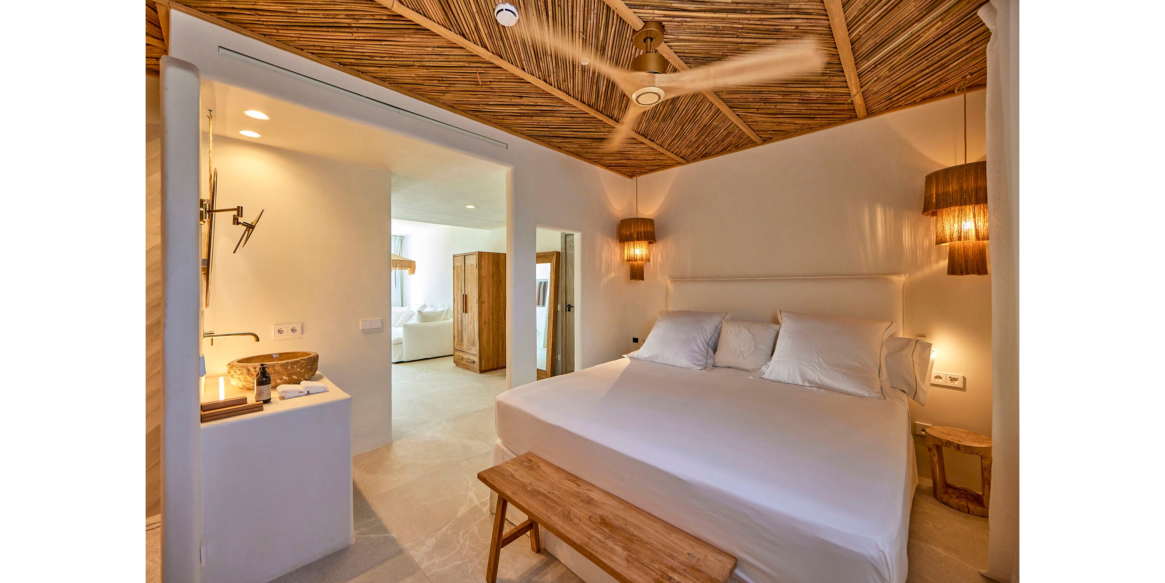 A contemporary bedroom features a large bed with crisp white linens and a simple wooden bench at the foot. The space has a warm tropical feel, highlighted by a bamboo-style ceiling and matching pendant lights. An open doorway reveals an adjoining room with additional furniture and soft natural light. Neutral tones and minimal decor create a calm, inviting atmosphere suitable for a resort or vacation home.