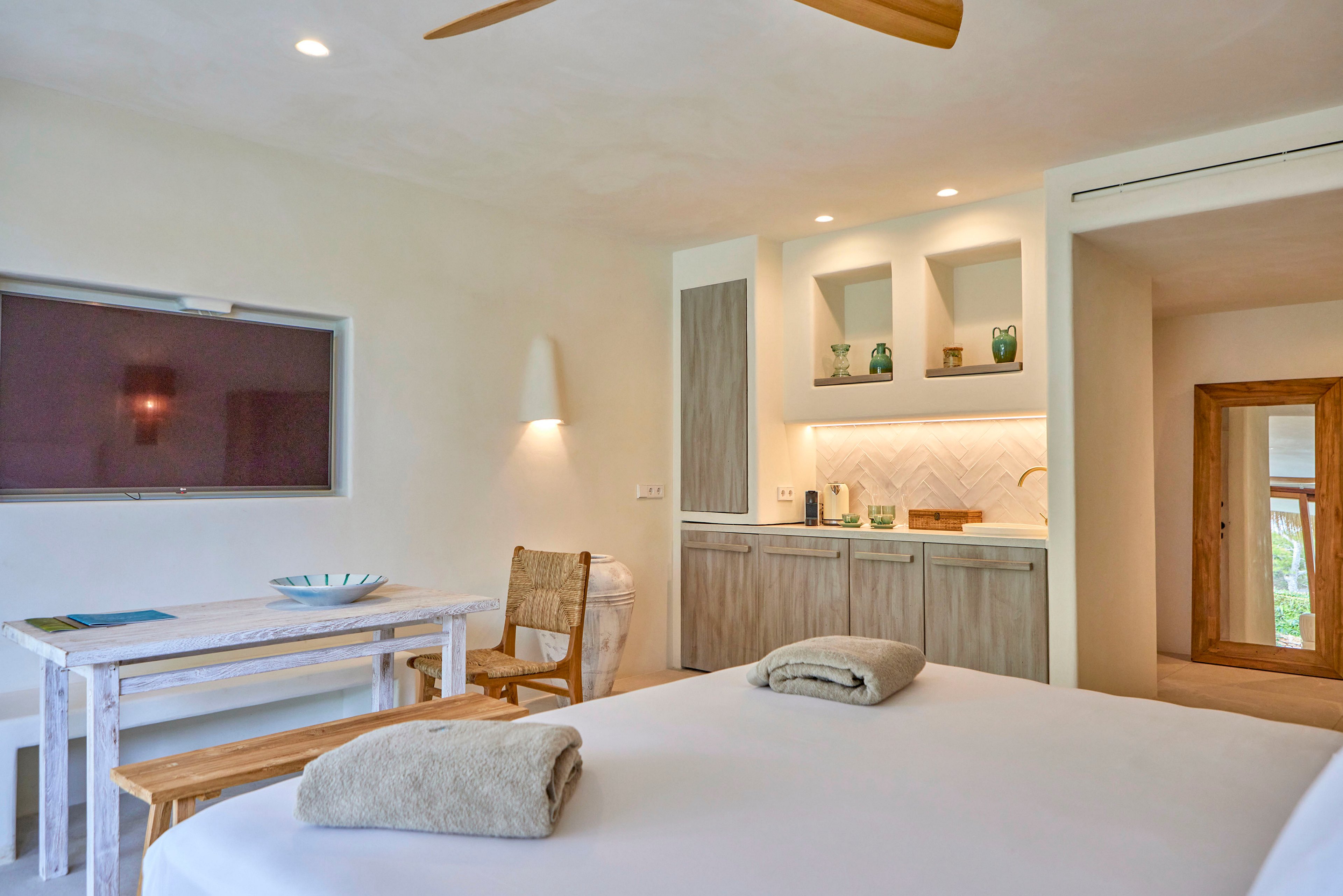 A bright, modern hotel room features a neatly made bed in the foreground with folded towels placed on top. In the background, a small kitchenette with open shelves, cabinets, and accent lighting adds functionality to the space. A wall-mounted flat-screen television faces a rustic white wooden table and bench, accompanied by a wicker chair. Warm neutral tones, wooden accents, and soft lighting create a calm, minimalist atmosphere.