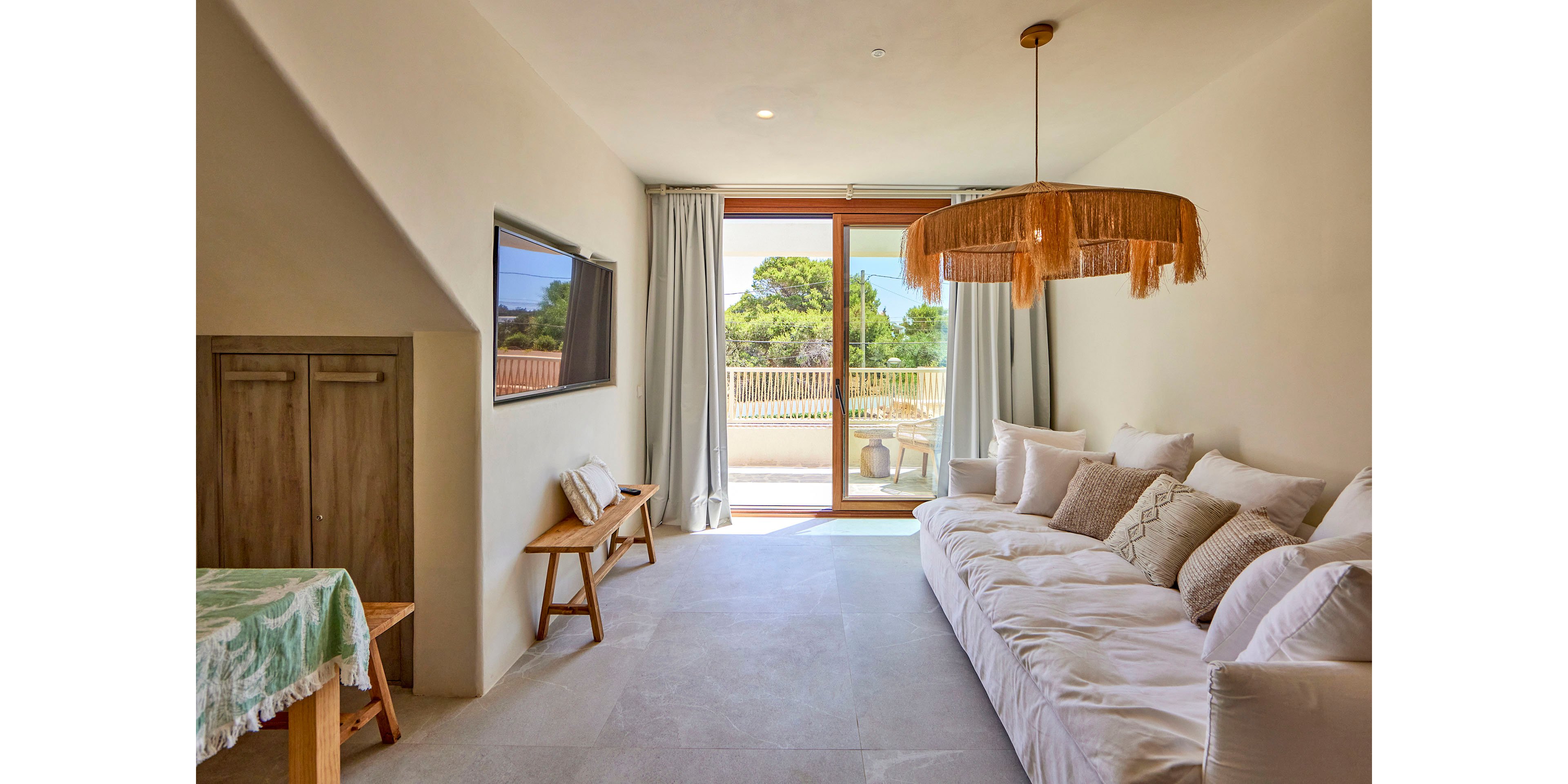 A bright, minimalist living room features a long white sofa lined with cushions along one wall. A flat-screen television is mounted opposite, with a wooden bench and small cabinet adding natural accents. Large glass doors with light curtains open to a sunny terrace and greenery outside. A woven pendant light and neutral tones create a relaxed, coastal atmosphere.