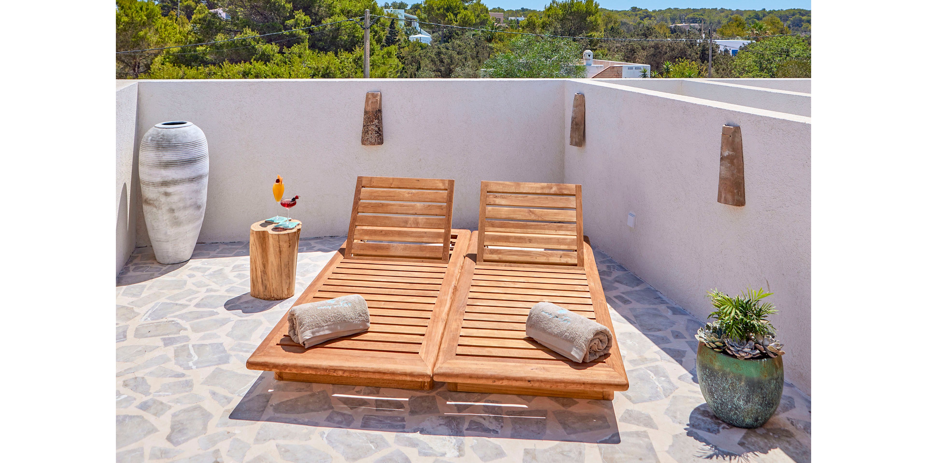 Two wooden sun loungers with rolled towels are placed on a stone-tiled rooftop terrace. A small wooden side table holds a colorful drink beside a tall ceramic vase. A potted plant in a blue-green pot adds greenery against the light-colored walls. Trees and a clear sky are visible beyond the terrace, creating a bright and relaxing outdoor setting.