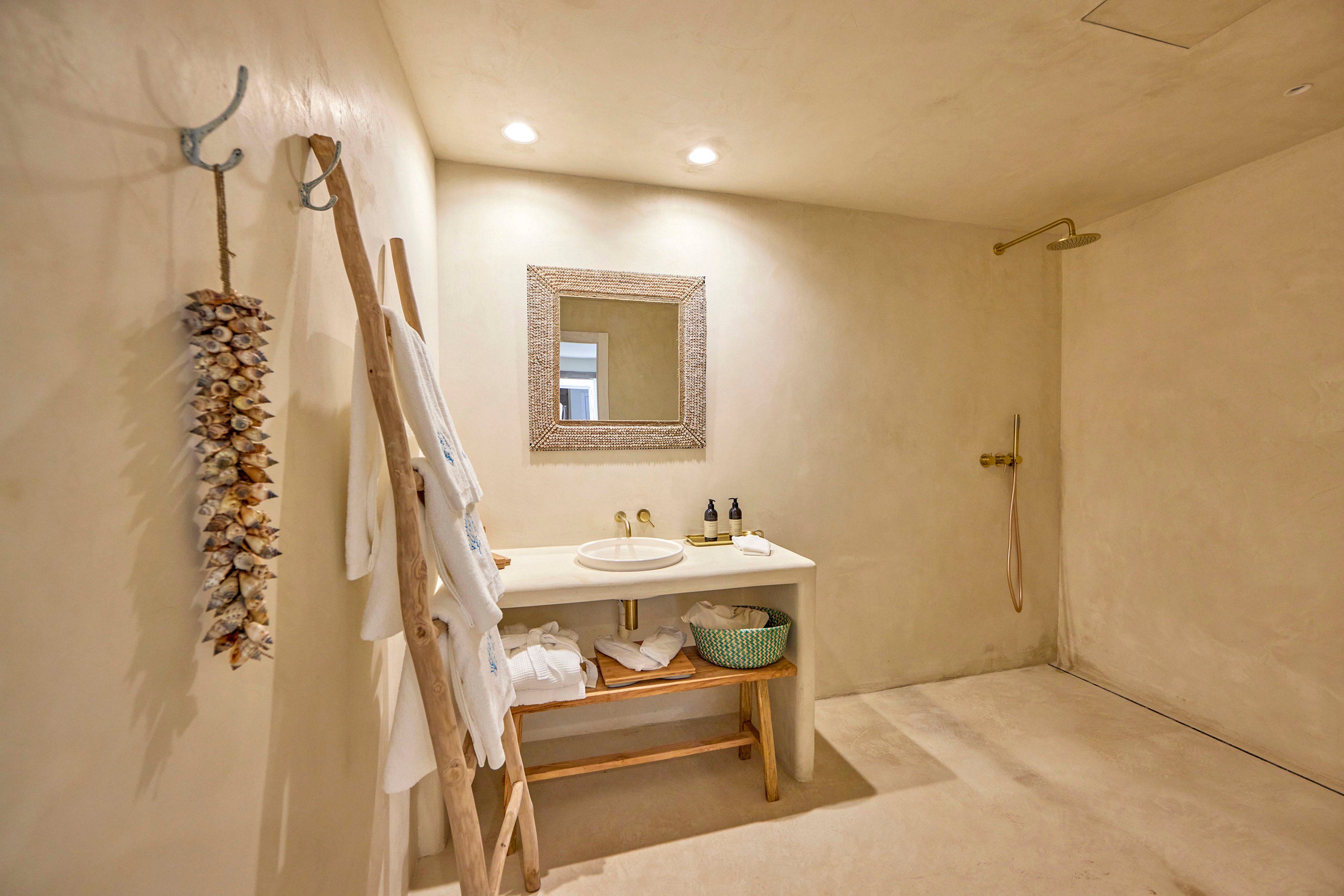 A spacious minimalist bathroom features a light beige color palette and smooth plaster walls. A simple wooden vanity with an inset sink, mirror, and toiletries sits against the wall, accompanied by a woven basket underneath. On the left, a wooden ladder holds white towels and robes, while decorative hanging shells add a coastal touch. A walk-in shower with a wall-mounted showerhead is visible on the right, illuminated by recessed ceiling lights.