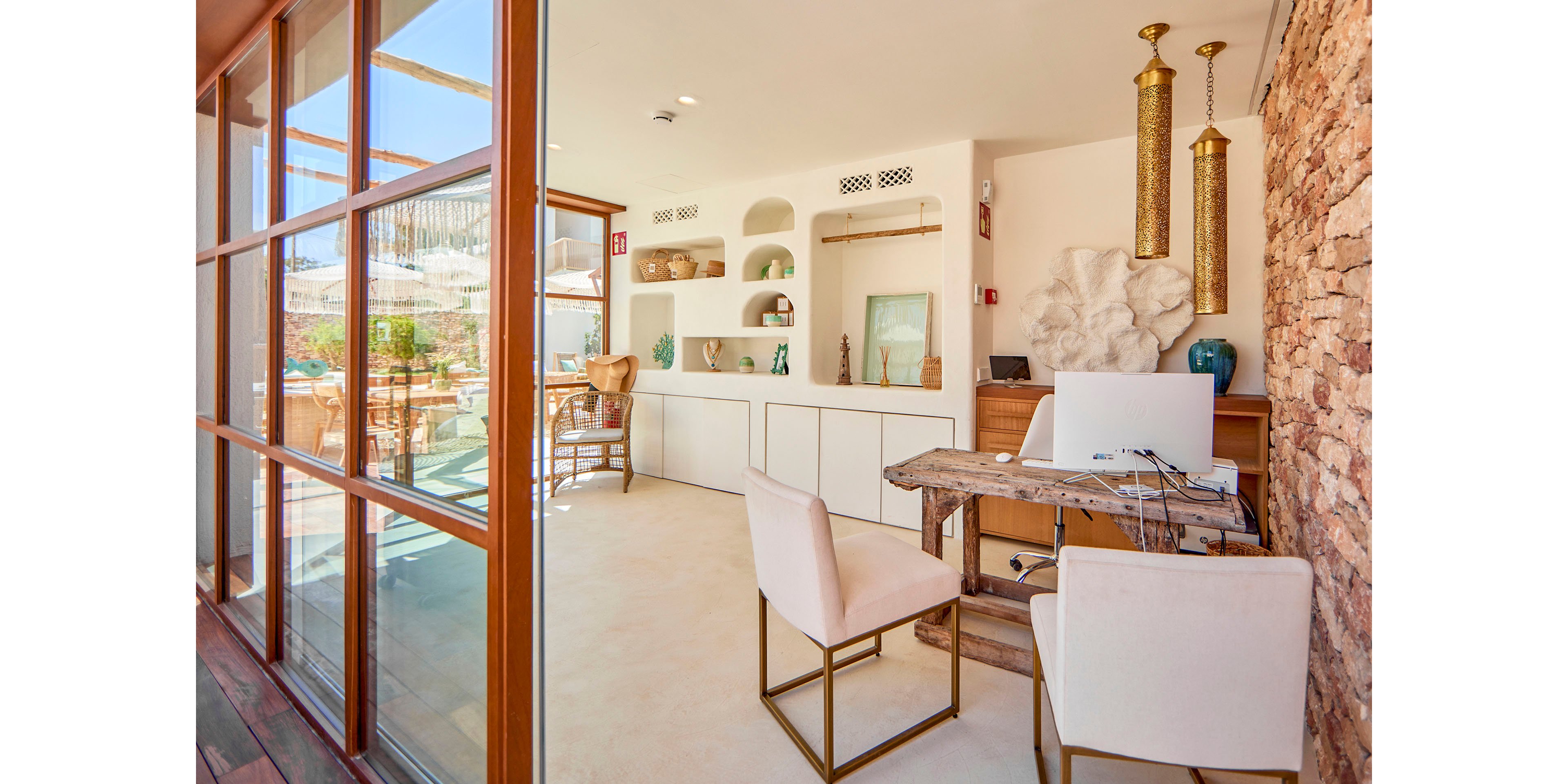 A sunlit office space features a rustic wooden desk and two modern upholstered chairs. Built-in white shelves display vases, bowls, and coastal-inspired decor, including coral and framed art. Large glass doors open to an outdoor terrace, filling the room with natural light and views of the surrounding landscape. Warm neutral tones, stone walls, and brass hanging lights create a relaxed, Mediterranean atmosphere.