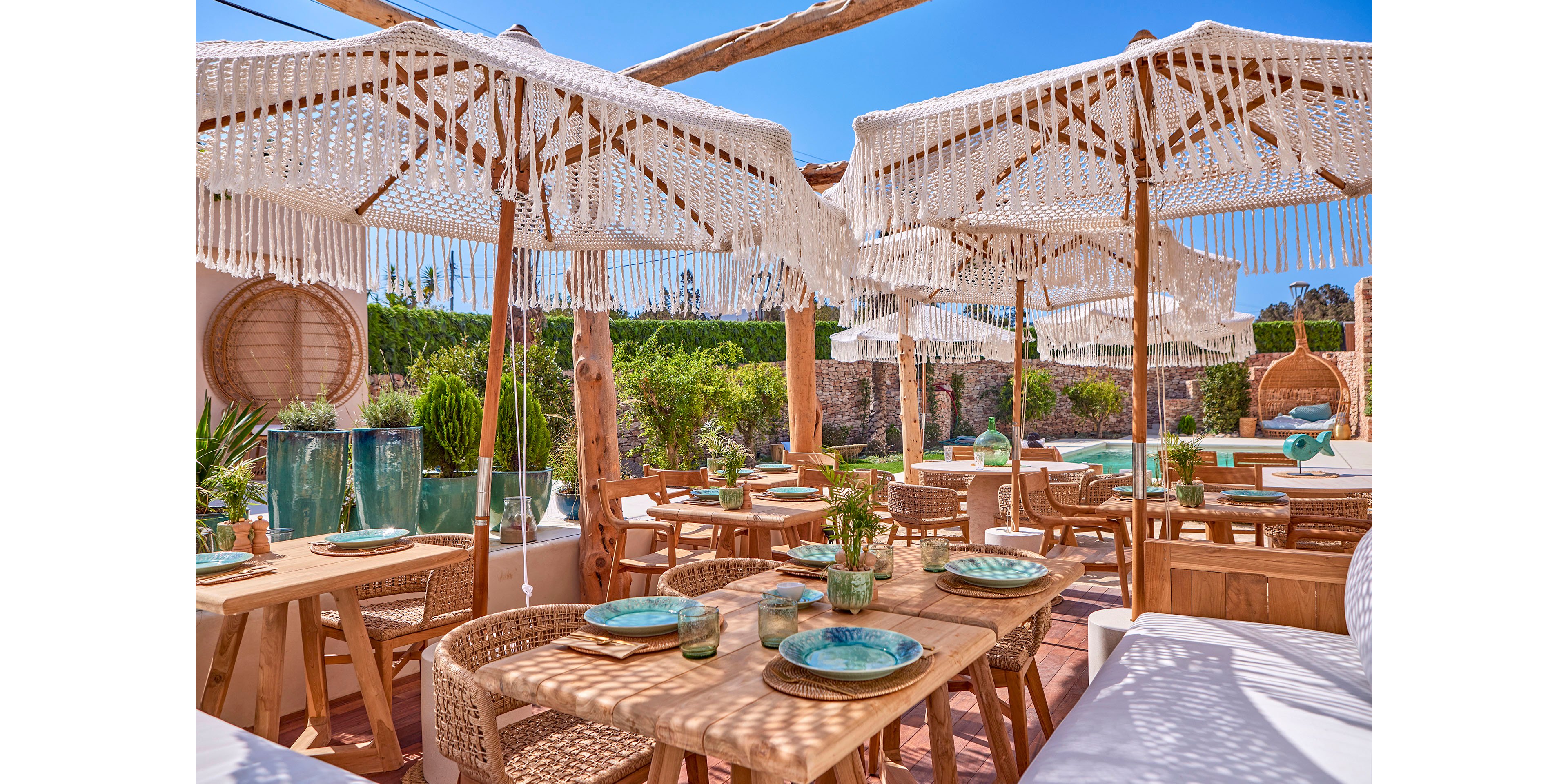 An open-air restaurant features wooden tables set with turquoise plates and glassware under fringed, woven parasols. Natural wicker chairs and benches create a relaxed, coastal atmosphere around the dining area. Lush greenery and a pool in the background enhance the resort-like setting. The bright blue sky and warm sunlight emphasize the summery, Mediterranean vibe.