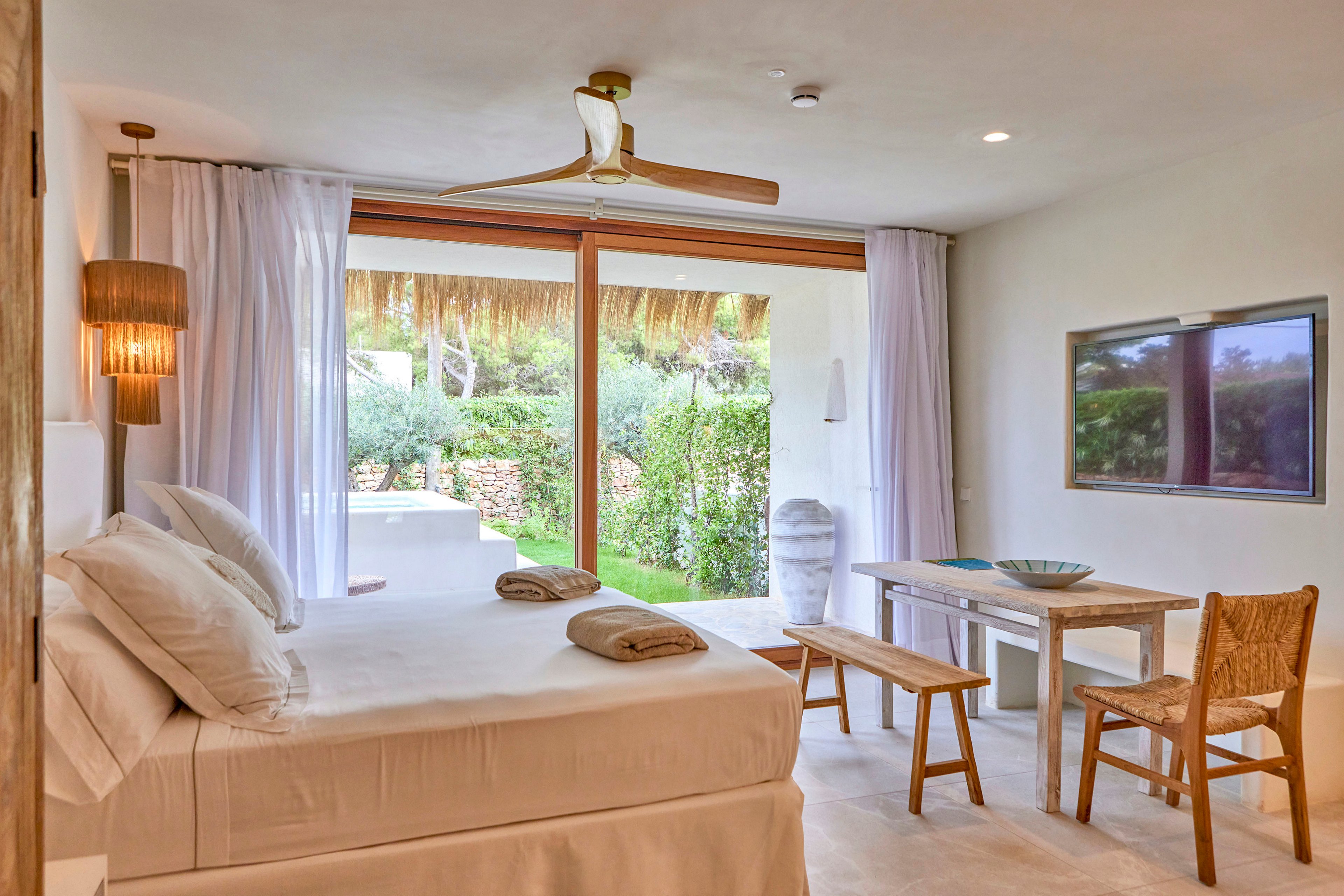 A serene, minimalist bedroom features a neatly made double bed with white linens and folded towels. Large sliding glass doors open to a lush green garden, filling the room with natural light. A ceiling fan, woven light fixtures, and natural wood furniture add warmth and texture to the neutral palette. A wall-mounted television and simple desk area complete the modern, tranquil interior.