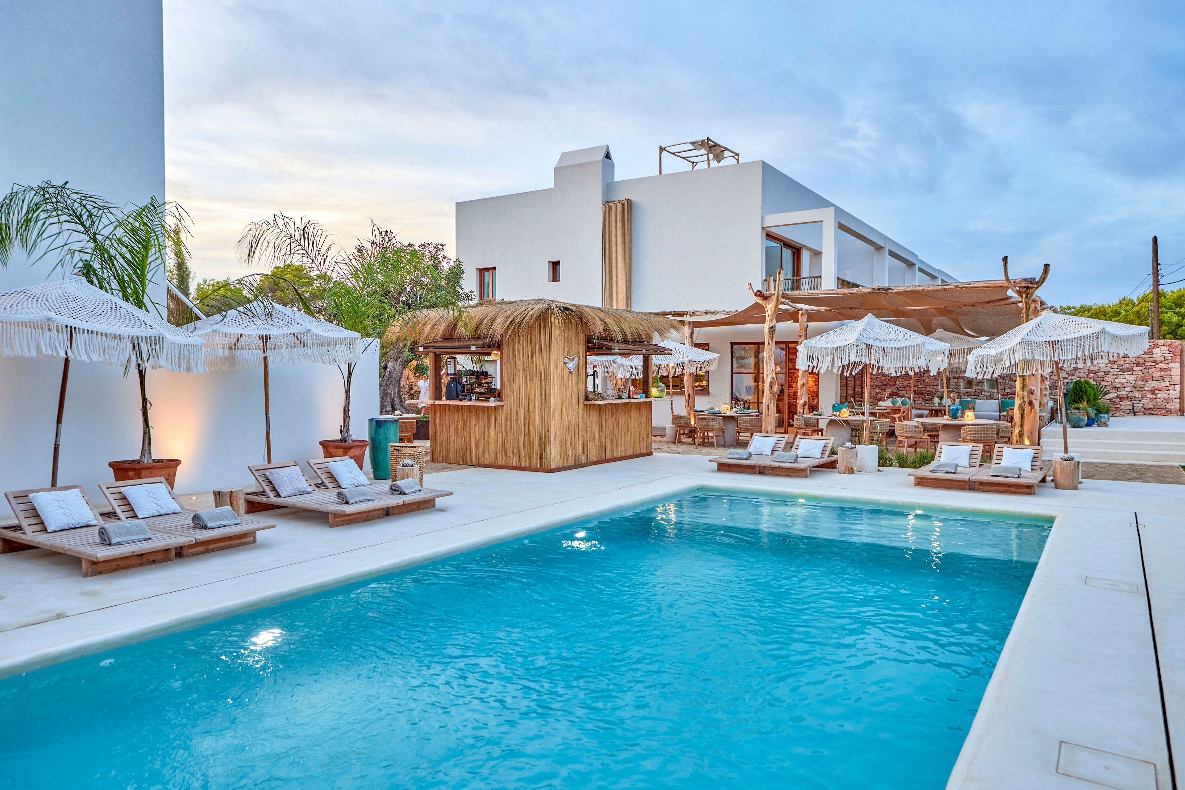 A modern outdoor swimming pool sits at the center of a stylish boutique hotel courtyard. Wooden sun loungers with white cushions and fringed parasols line the poolside, creating a relaxed resort atmosphere. A thatched-roof bar and shaded seating area provide a tropical touch against the clean white architecture. Soft evening light and a calm blue sky enhance the tranquil, vacation-like setting.