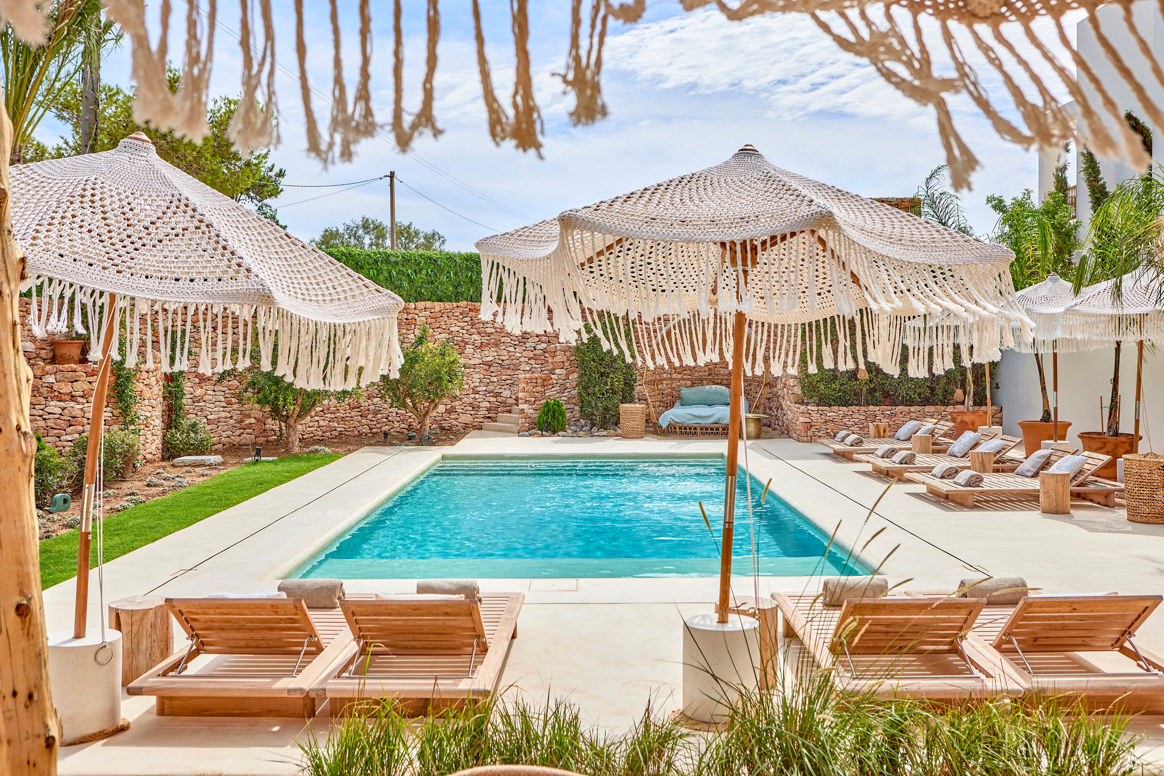 A bright outdoor swimming pool is surrounded by wooden loungers and bohemian-style parasols with macramé fringe. The scene is set in a private courtyard with stone walls, green hedges, and manicured grass. Cushioned daybeds and side tables line the far side of the pool, creating a relaxed resort atmosphere. Clear blue water and soft neutral tones emphasize a calm, summery mood.