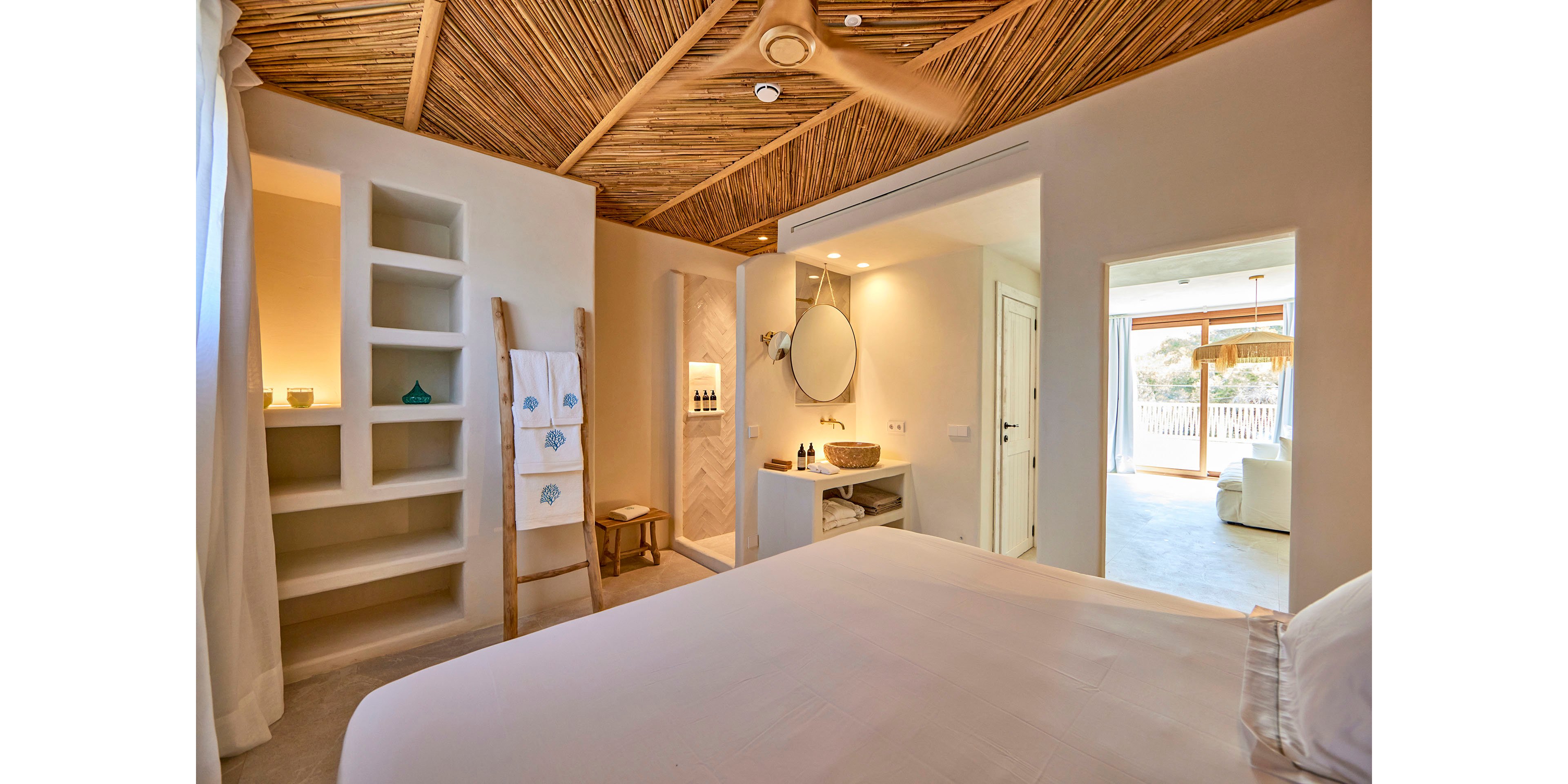 A serene bedroom features a large neatly made bed facing an open doorway filled with sunlight. The space has a natural wood ceiling with a ceiling fan and recessed lighting, creating a warm, coastal atmosphere. Built-in shelves, a ladder with towels, and a compact vanity area with a round mirror add functional details. Beyond the doorway, an adjoining sitting area and outdoor view enhance the airy, resort-like feel.