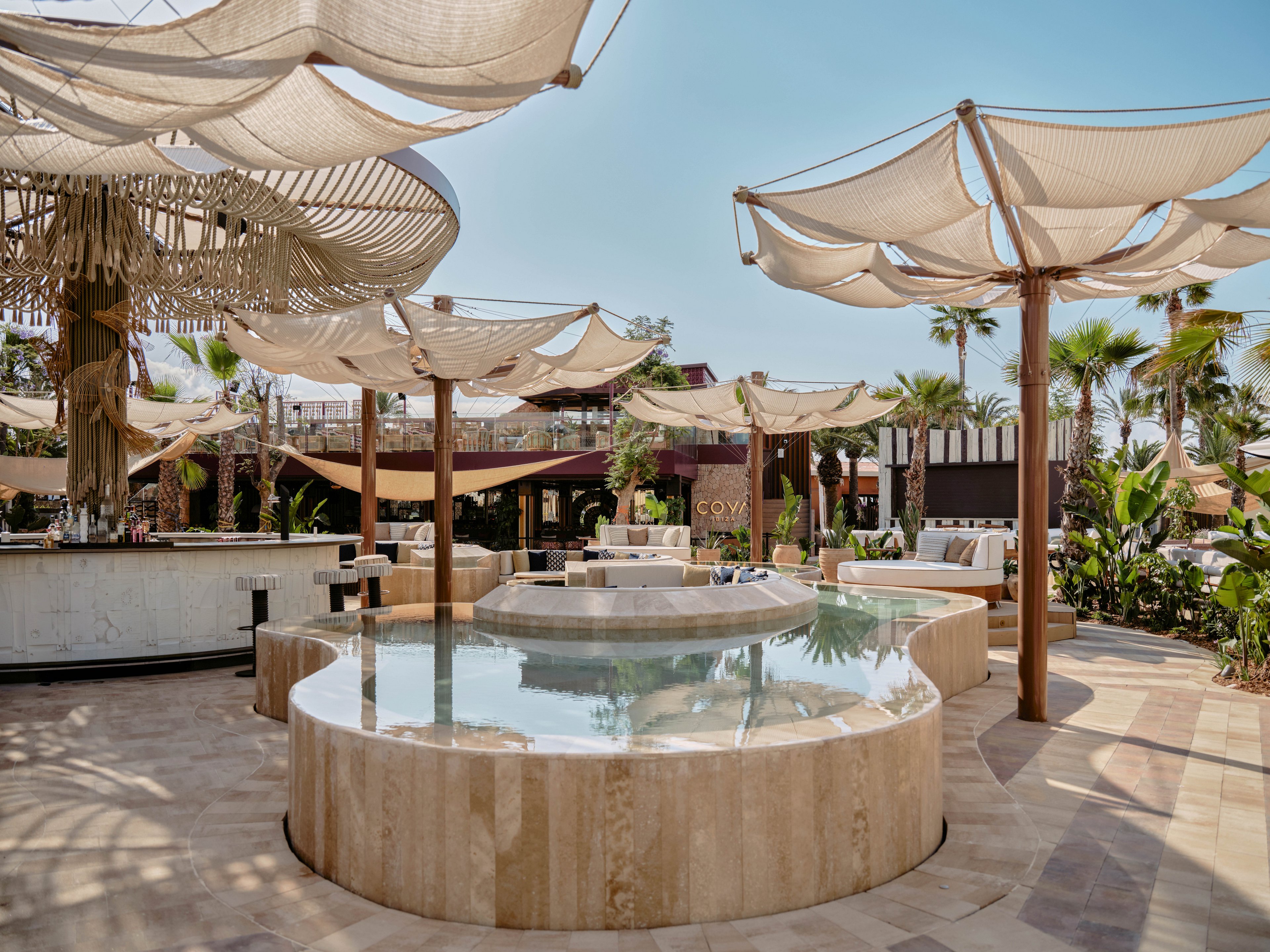 An upscale outdoor lounge features a circular shallow pool surrounded by stone seating. Fabric canopies are stretched above on wooden poles, creating sculptural shade structures. The setting includes cushioned sofas, palm trees, and tropical greenery, suggesting a resort or beach club atmosphere. Bright daylight and neutral tones give the scene a relaxed, sophisticated mood.