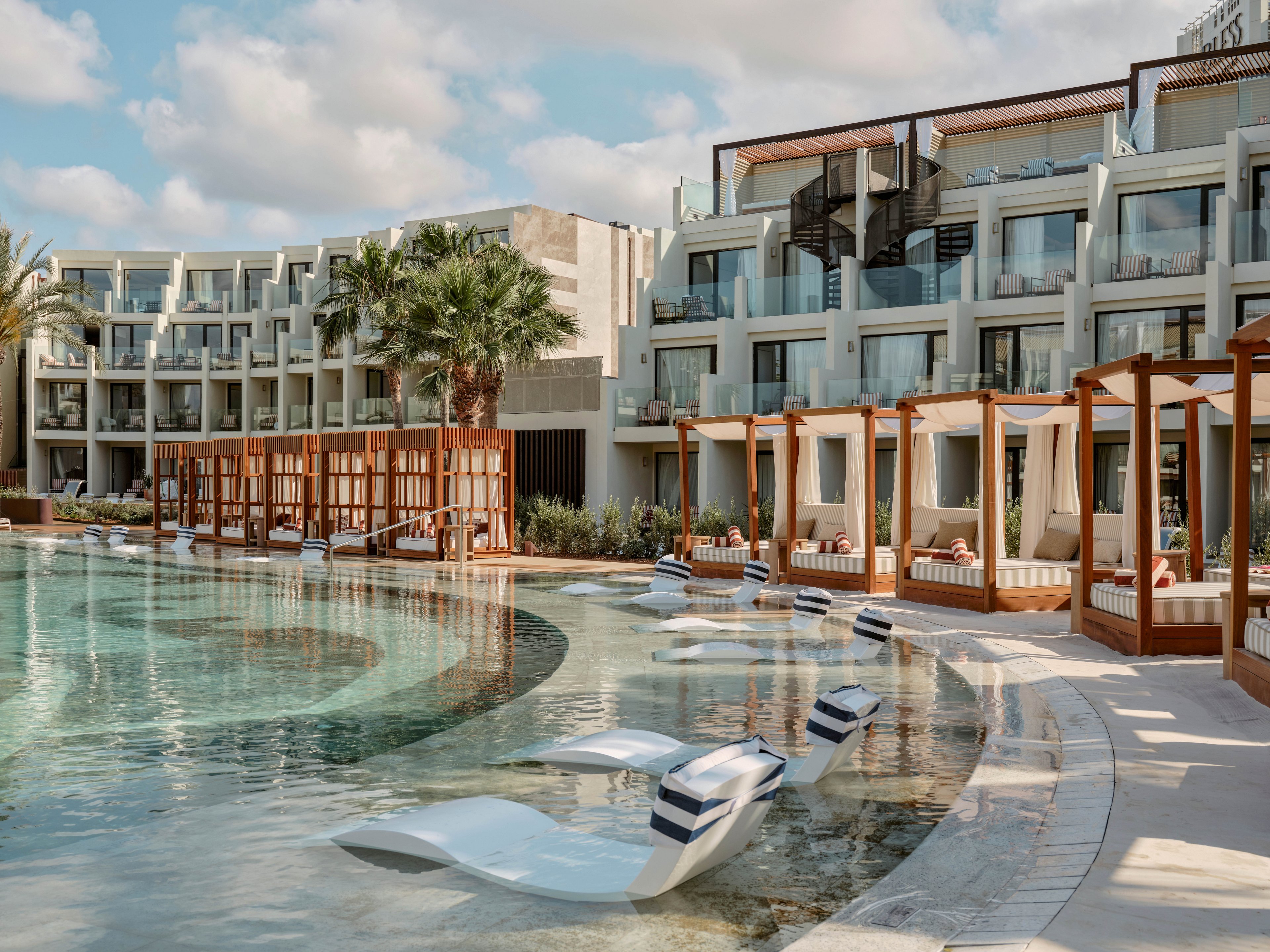 A modern resort features a large curved swimming pool lined with in-water loungers and striped cushions. Wooden cabanas with seating areas and daybeds border the pool, offering shaded relaxation spaces. The multi-story hotel building with balconies rises in the background under a partly cloudy sky. Palm trees and warm neutral tones create a sophisticated, tranquil vacation atmosphere.