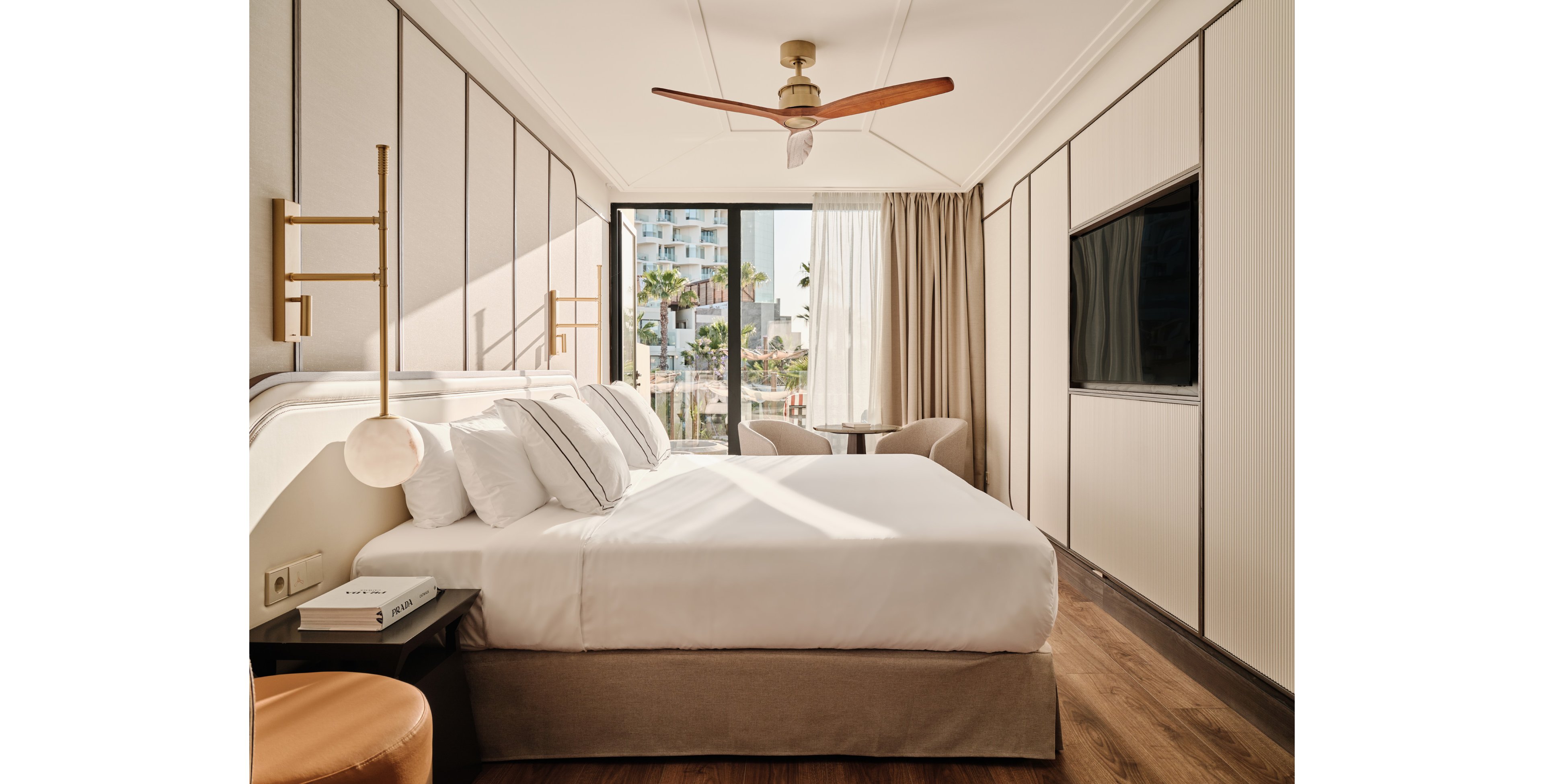 A contemporary hotel bedroom features a neatly made double bed with white linens and multiple pillows. The room includes a large wall-mounted television, a ceiling fan, and minimalist bedside lighting. Floor-to-ceiling glass doors open to a balcony with outdoor seating and views of nearby buildings and palm trees. Warm wood flooring and neutral tones create a calm, inviting atmosphere.