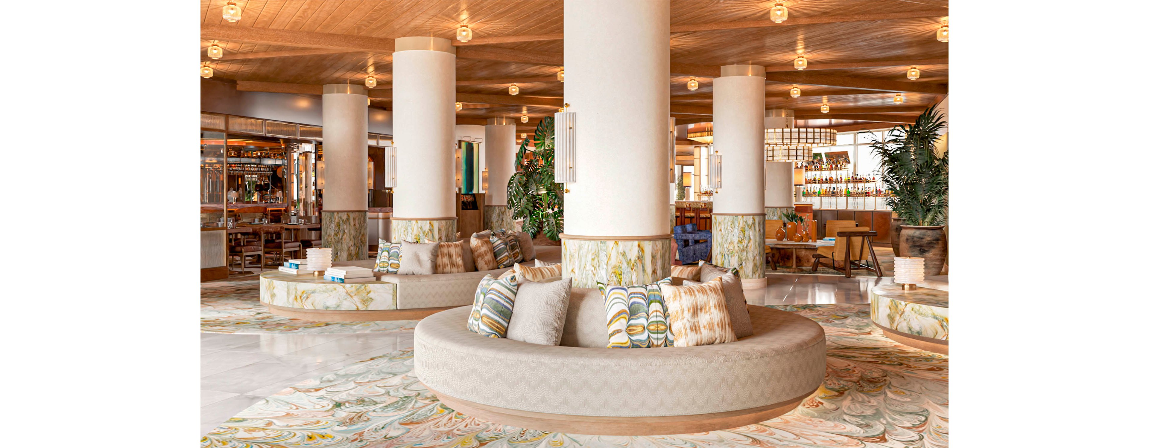 Spacious hotel lobby interior featuring large round sofas with patterned cushions around white columns. Warm wooden ceiling and soft ambient lighting create an inviting atmosphere. Decorative plants, tiled column bases, and a patterned floor add texture and color. A bar area and seating are visible in the background, suggesting a relaxed social space.