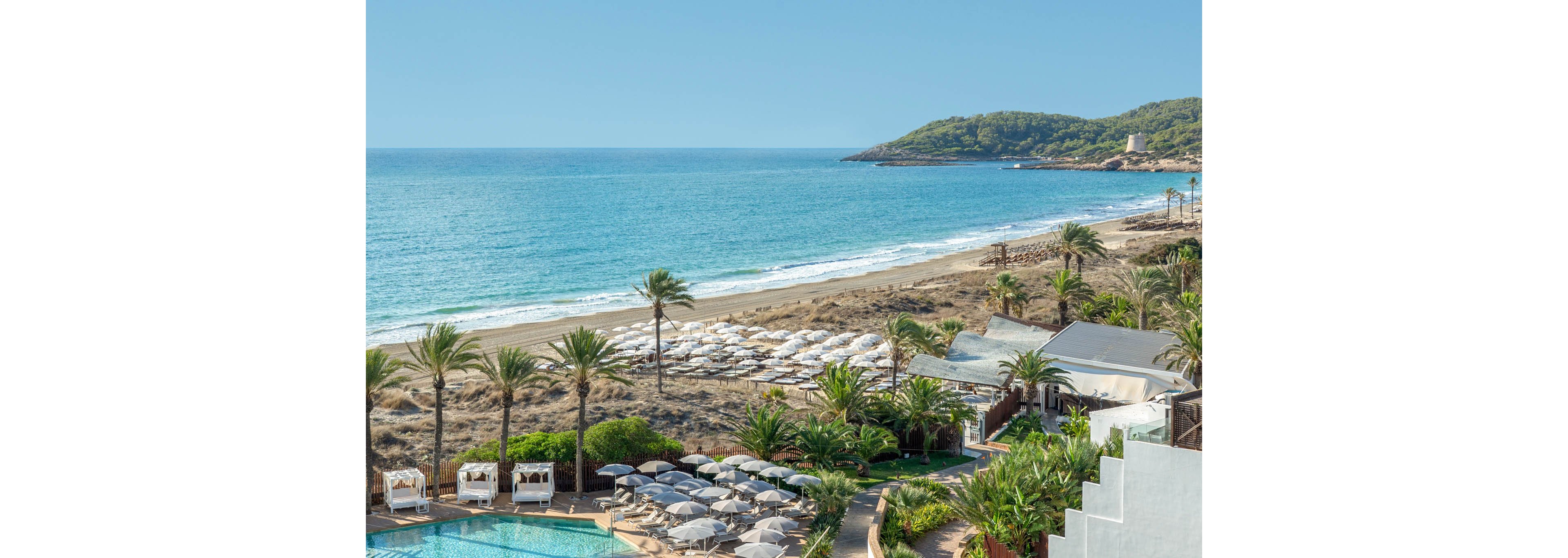 A coastal resort overlooks a sandy beach lined with rows of white sun loungers and umbrellas. In the foreground, a turquoise swimming pool is surrounded by palm trees and cushioned daybeds. The calm blue sea stretches to the horizon, with gentle waves meeting the shoreline. Green hills and a small white structure on the distant headland complete the serene vacation setting.