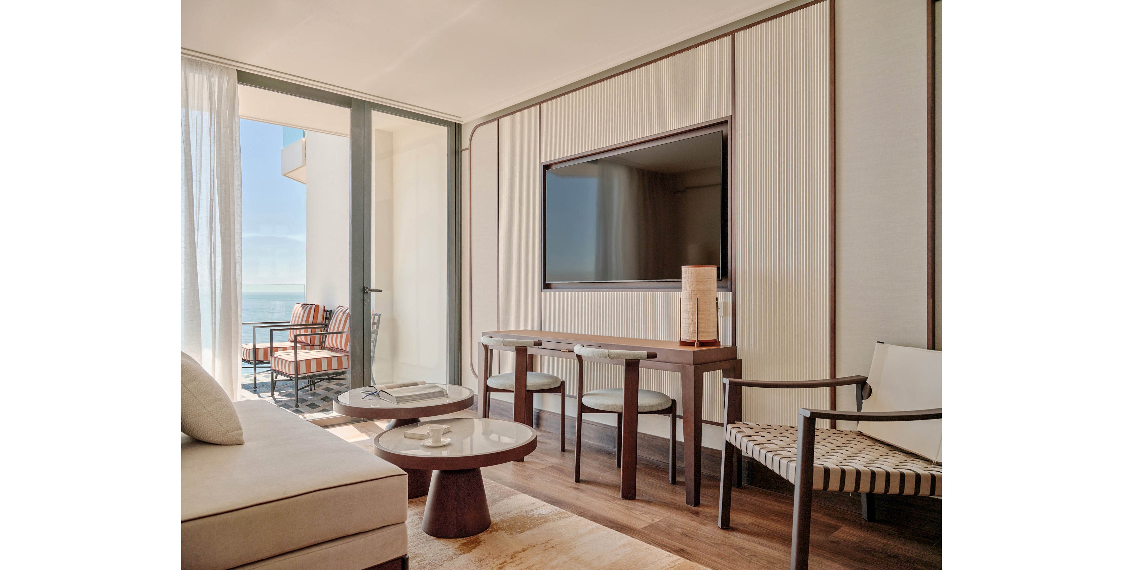 A contemporary hotel living area features a large wall-mounted television and minimalist wooden furniture. Light neutral tones dominate the space, with a sofa, armchair, and round coffee tables arranged for relaxation. Floor-to-ceiling glass doors open to a balcony with striped chairs and an ocean view. Soft natural light filters through sheer curtains, creating an airy and inviting atmosphere.