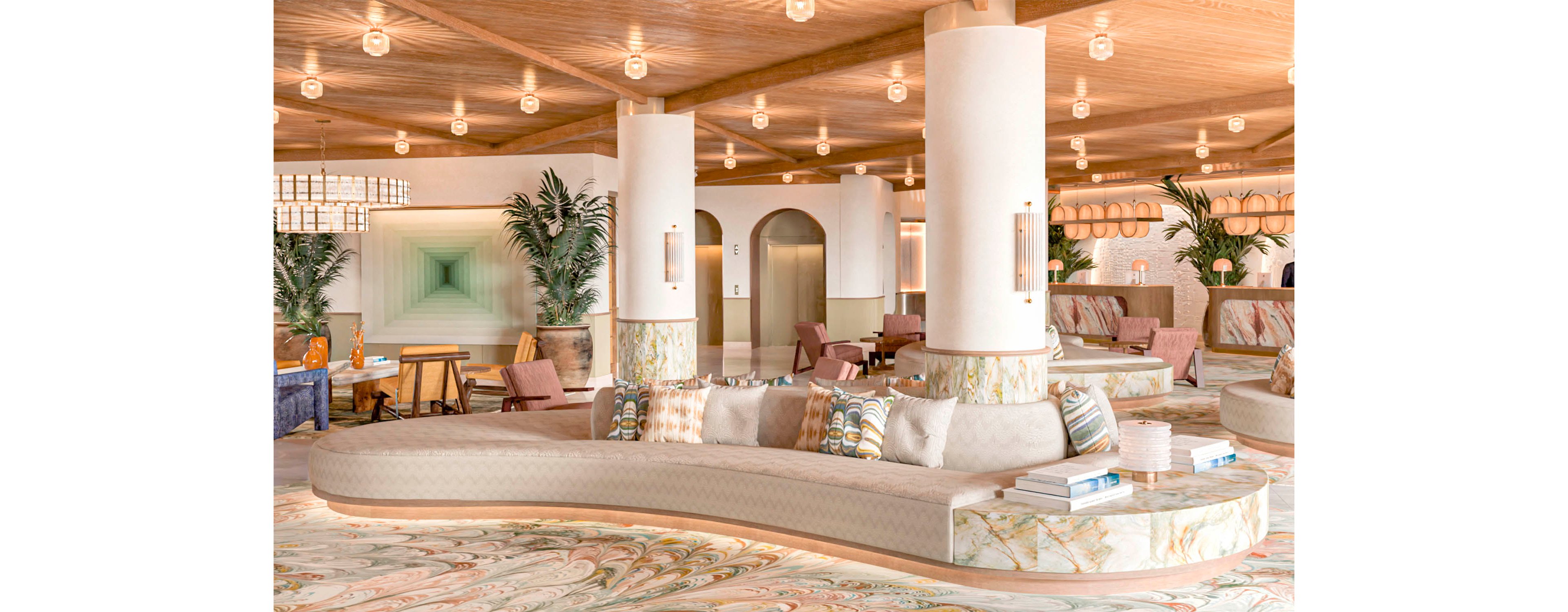 Spacious hotel lobby features a large curved sofa with patterned cushions and books arranged on marble side sections. The setting includes tall white columns, warm wooden ceiling, and soft ambient lighting from numerous pendant lights. Decorative plants, abstract wall art, and upholstered armchairs create a relaxed, upscale atmosphere. Overall, the space feels inviting, modern, and designed for socializing or quiet relaxation.